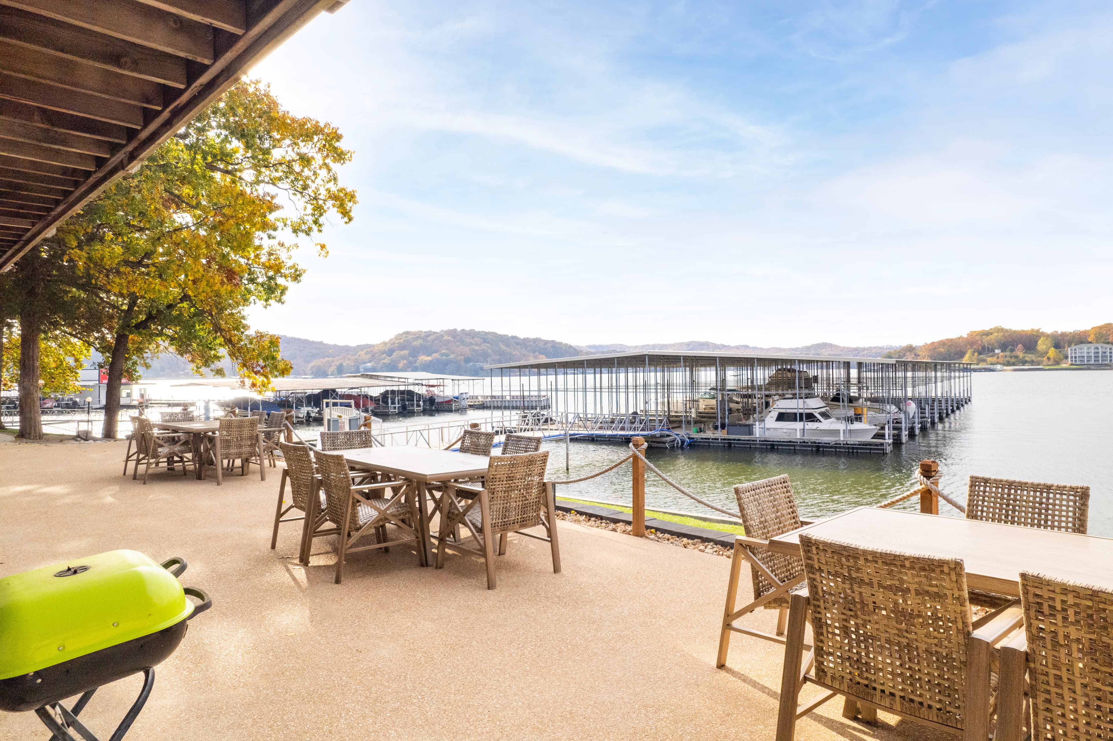 A lakeside patio with wooden tables overlooks a marina filled with boats under a clear blue sky.