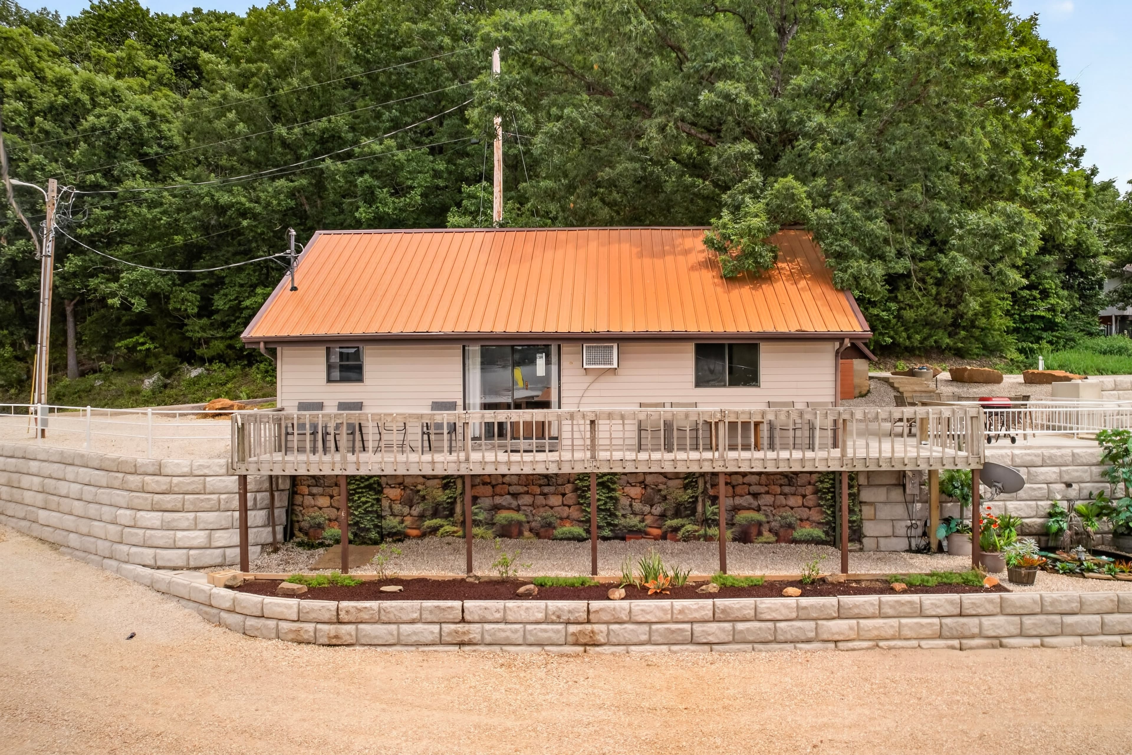A single-story house with a metal roof, surrounded by greenery and featuring a wooden deck.