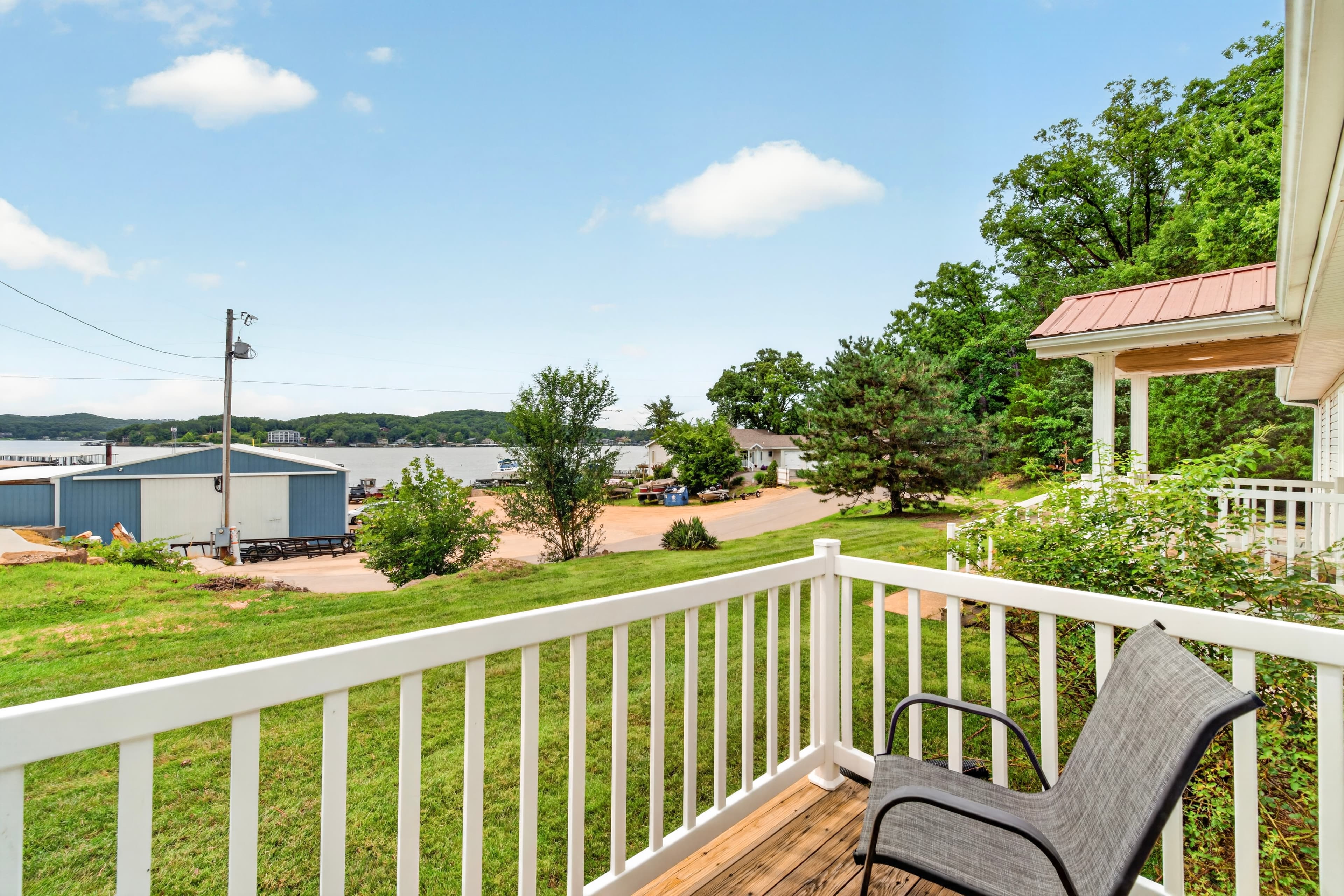 A view from a porch overlooking a grassy area, trees, and distant buildings by a lake under a blue sky.