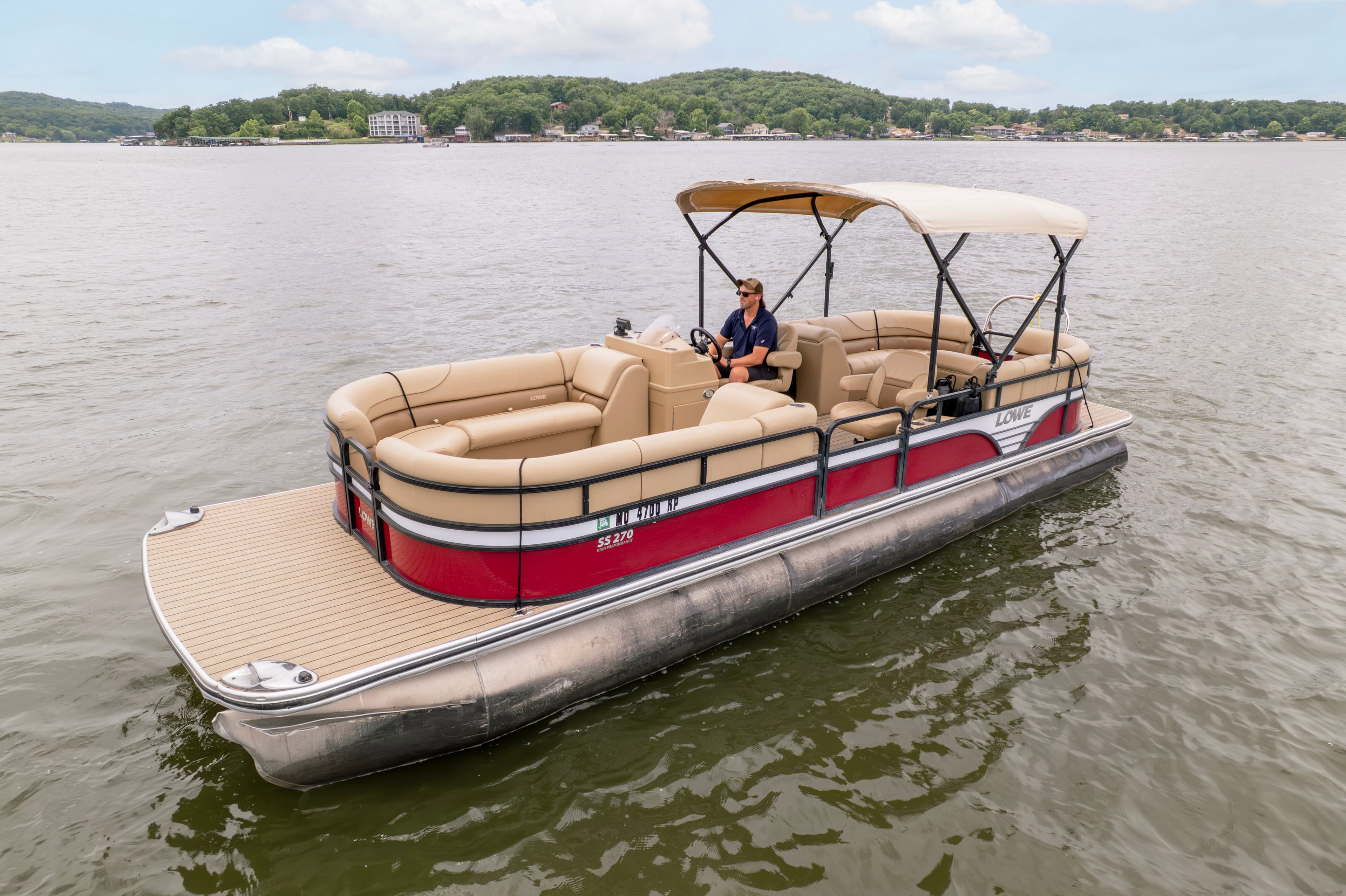 A person is steering a red and beige pontoon boat on a tranquil lake.