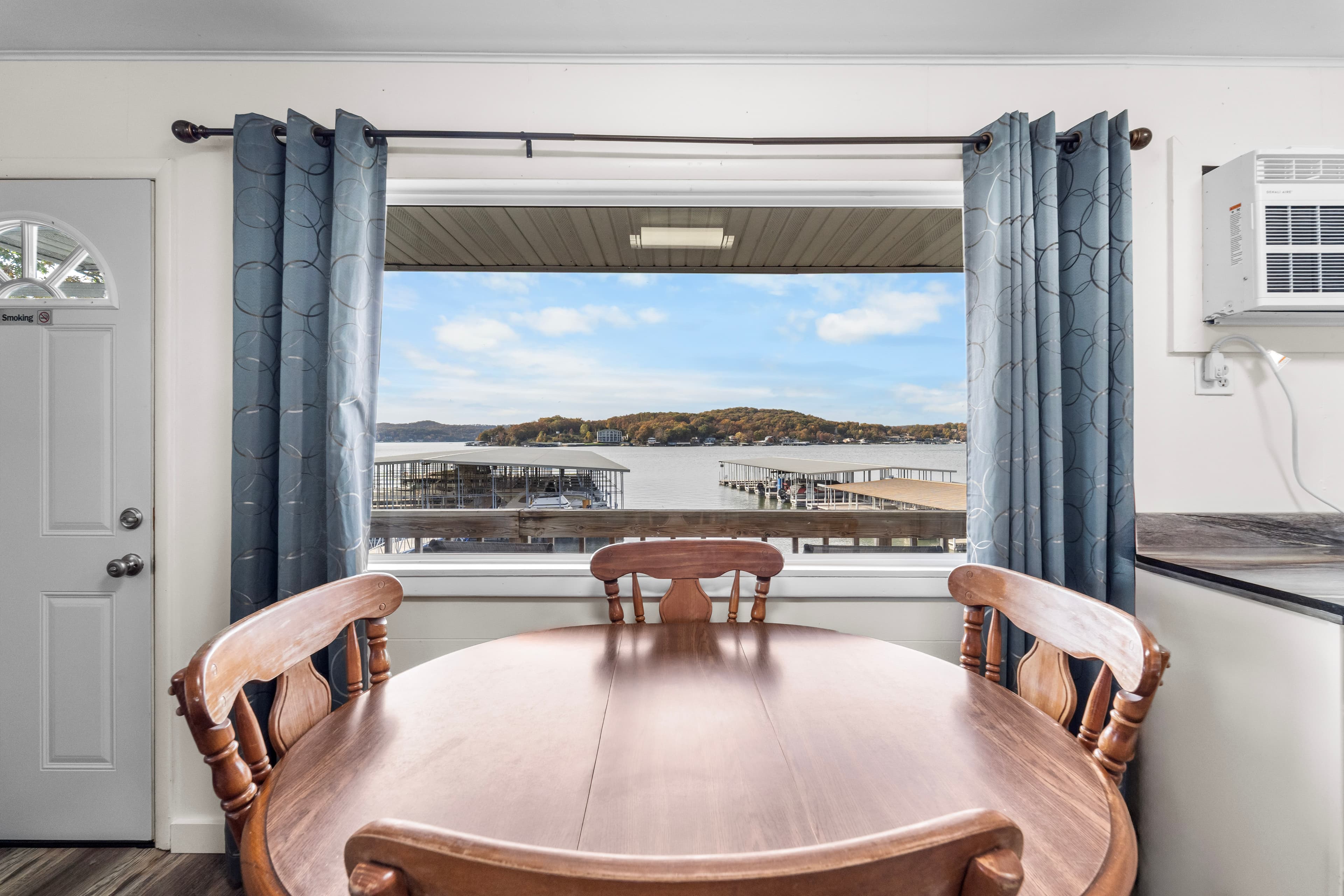 A wooden dining table sits in front of a large window with a view of a lake and shoreline.