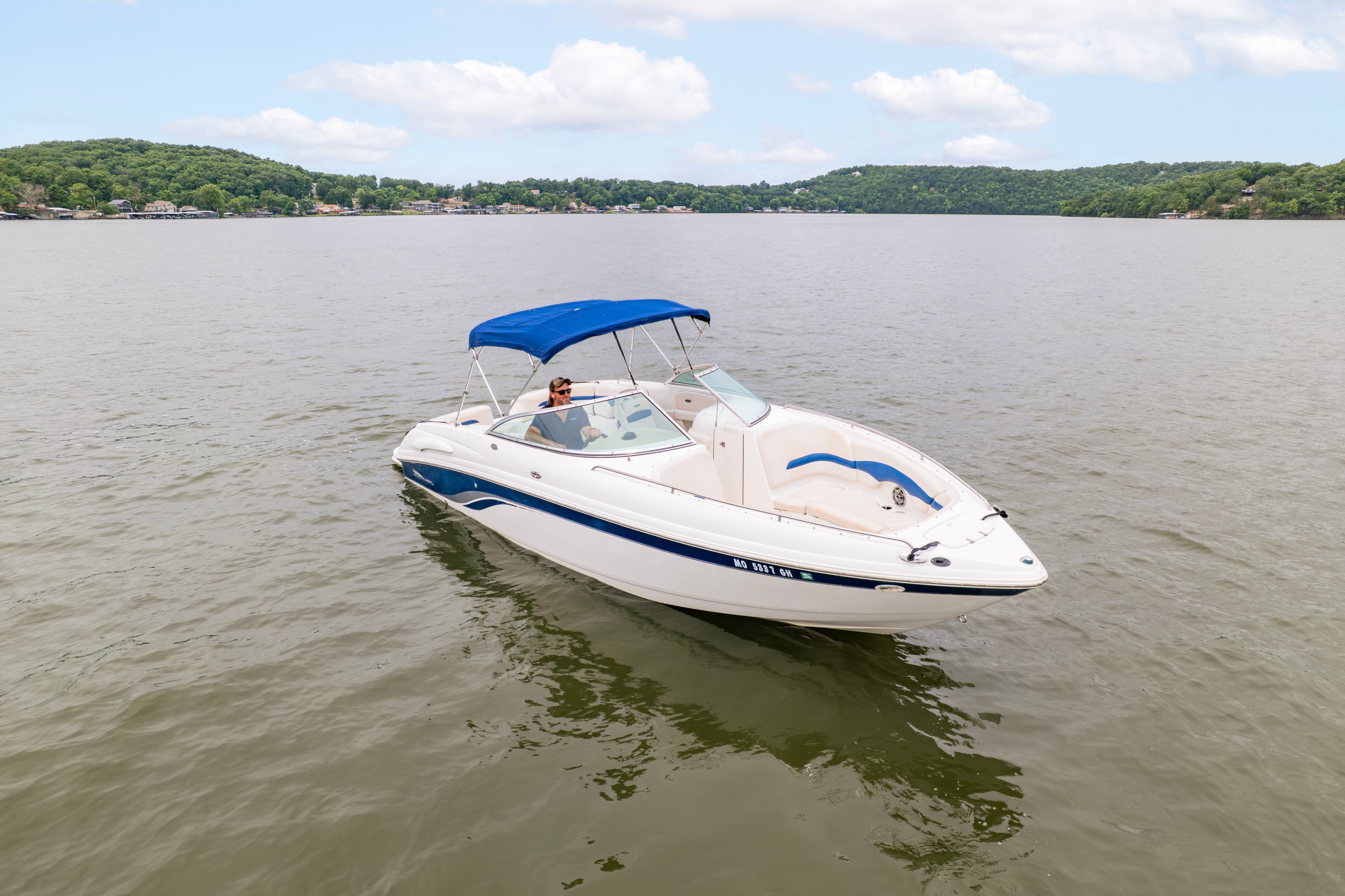 A white boat with a blue canopy is floating on calm water, surrounded by green hills under a partly cloudy sky.