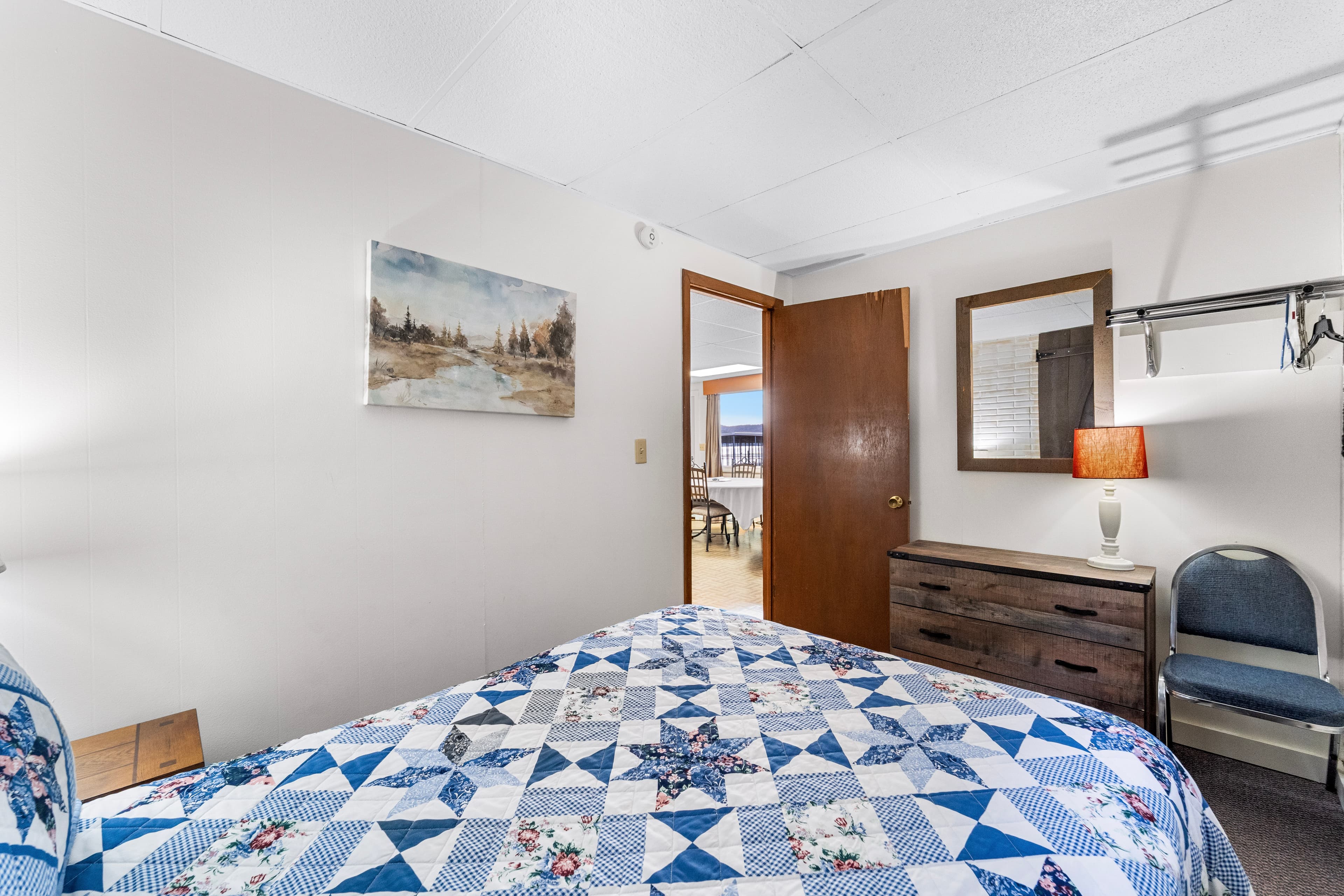A cozy bedroom featuring a patterned quilt, a wooden dresser, a mirror, and a door leading to another space.