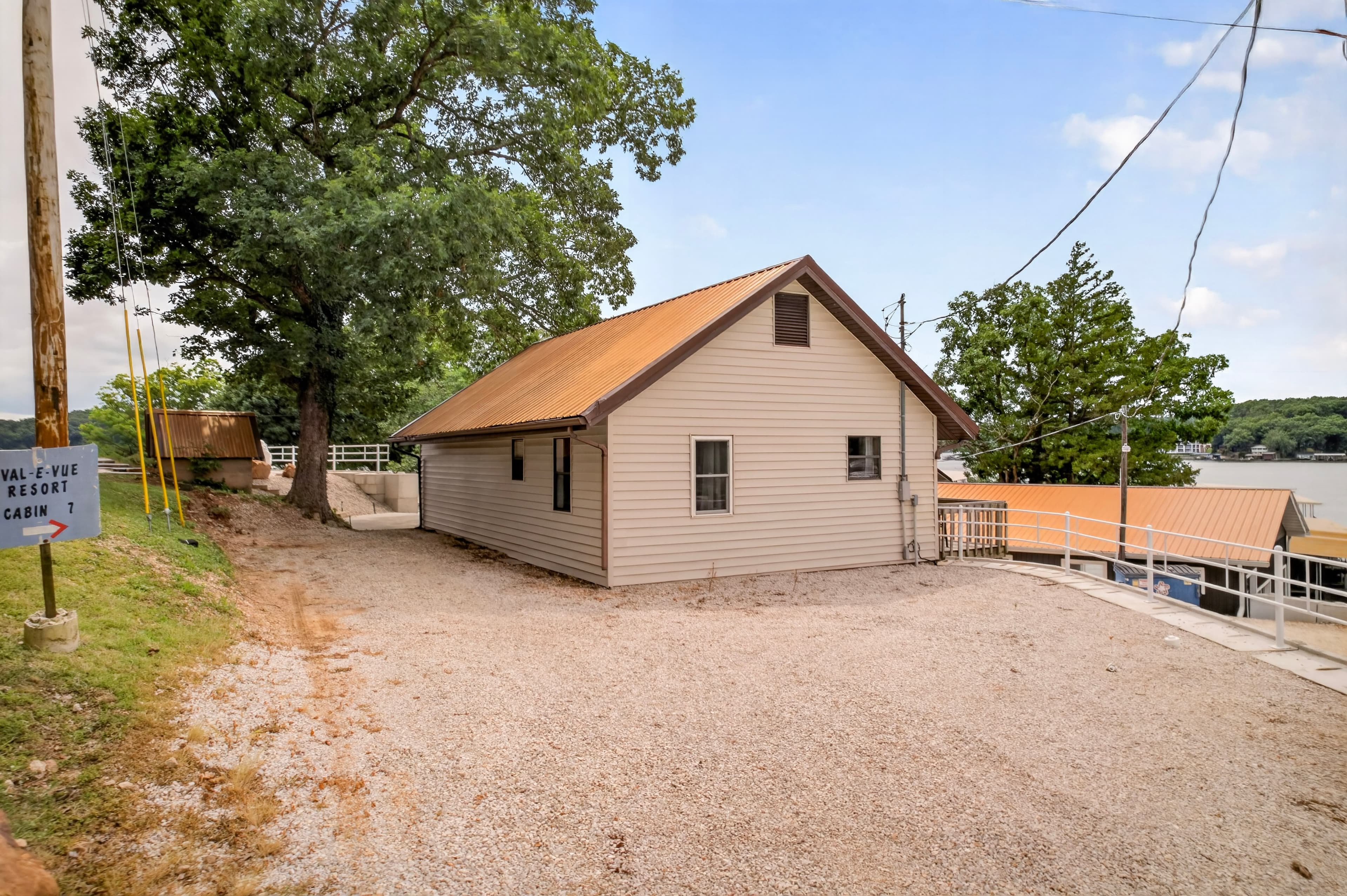 A small cabin with a metal roof is situated near a lake, surrounded by greenery and gravel paths.