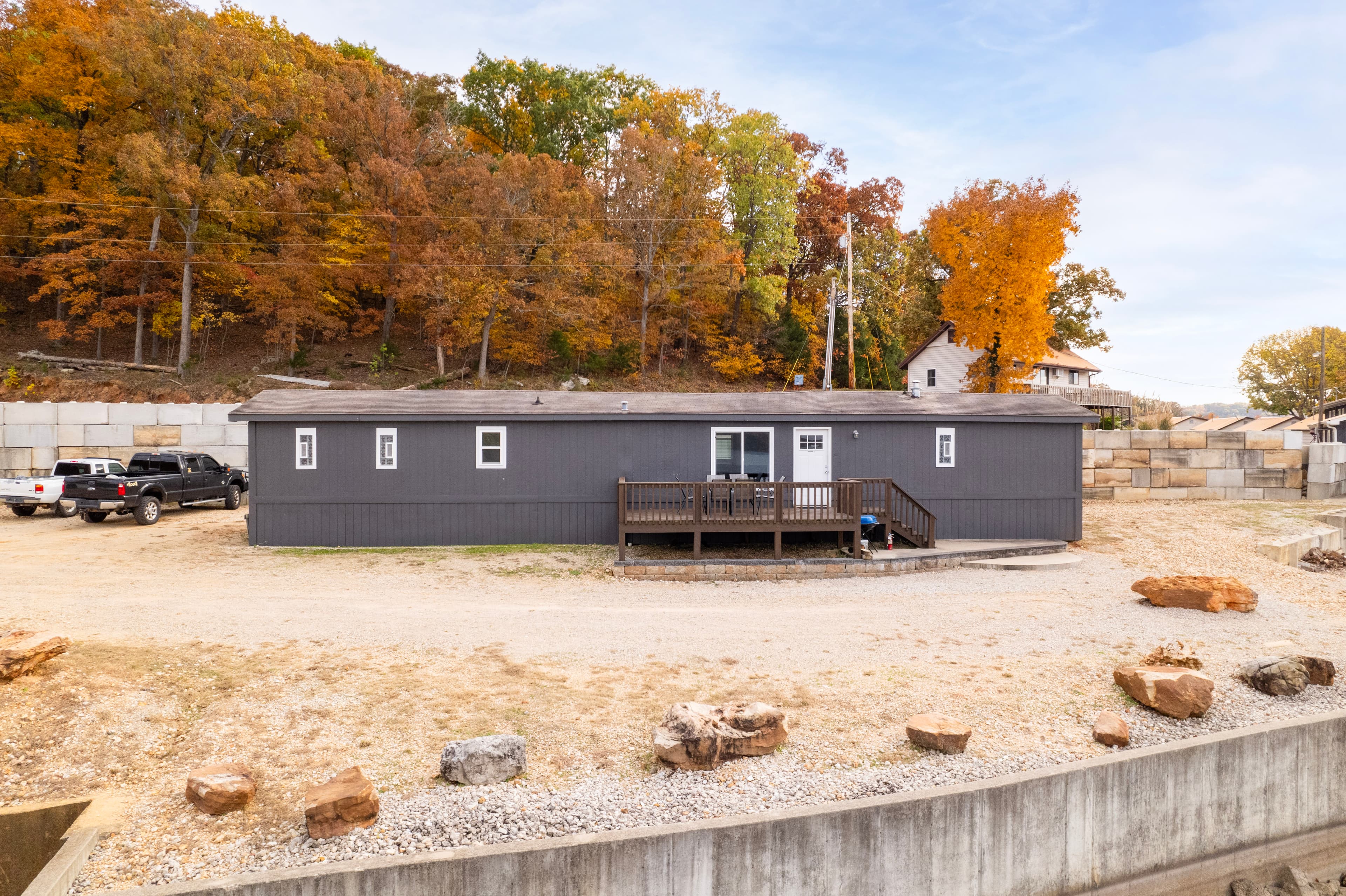 A gray modular home sits on a gravel lot surrounded by trees with autumn foliage.