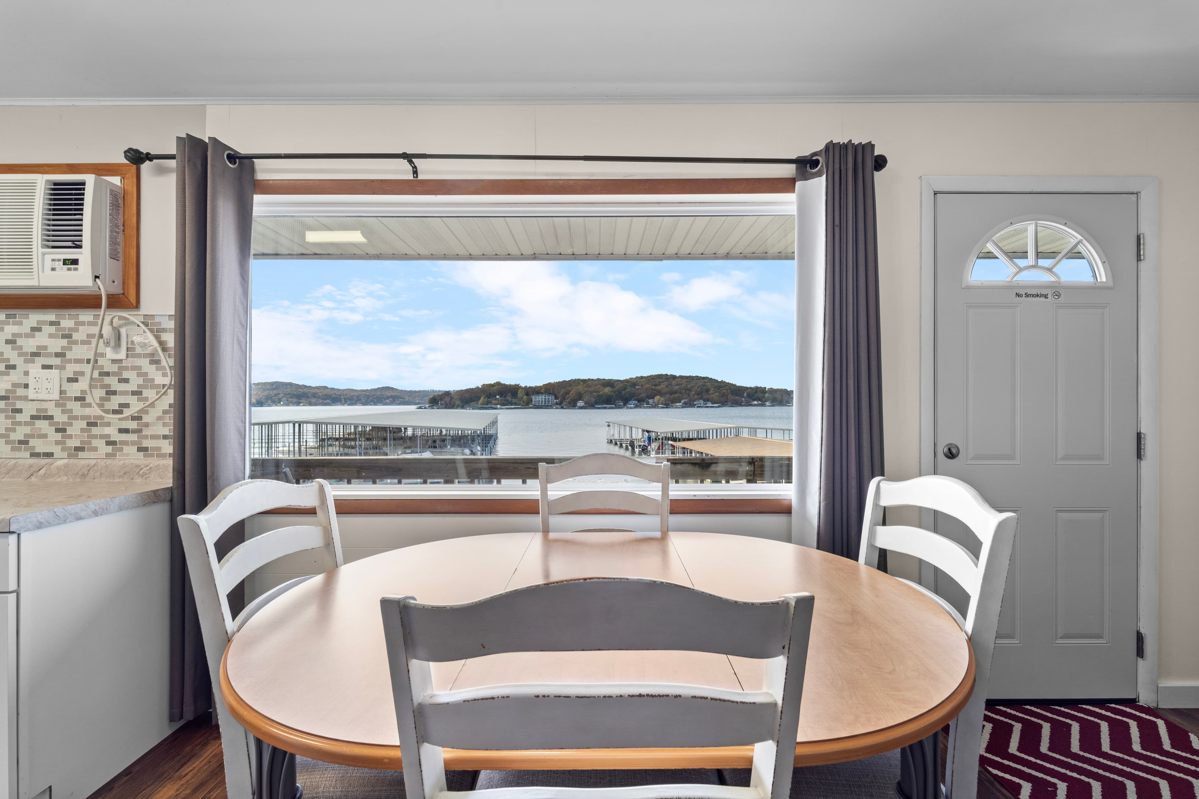 A dining table sits in front of a large window overlooking a scenic view of hills and water.