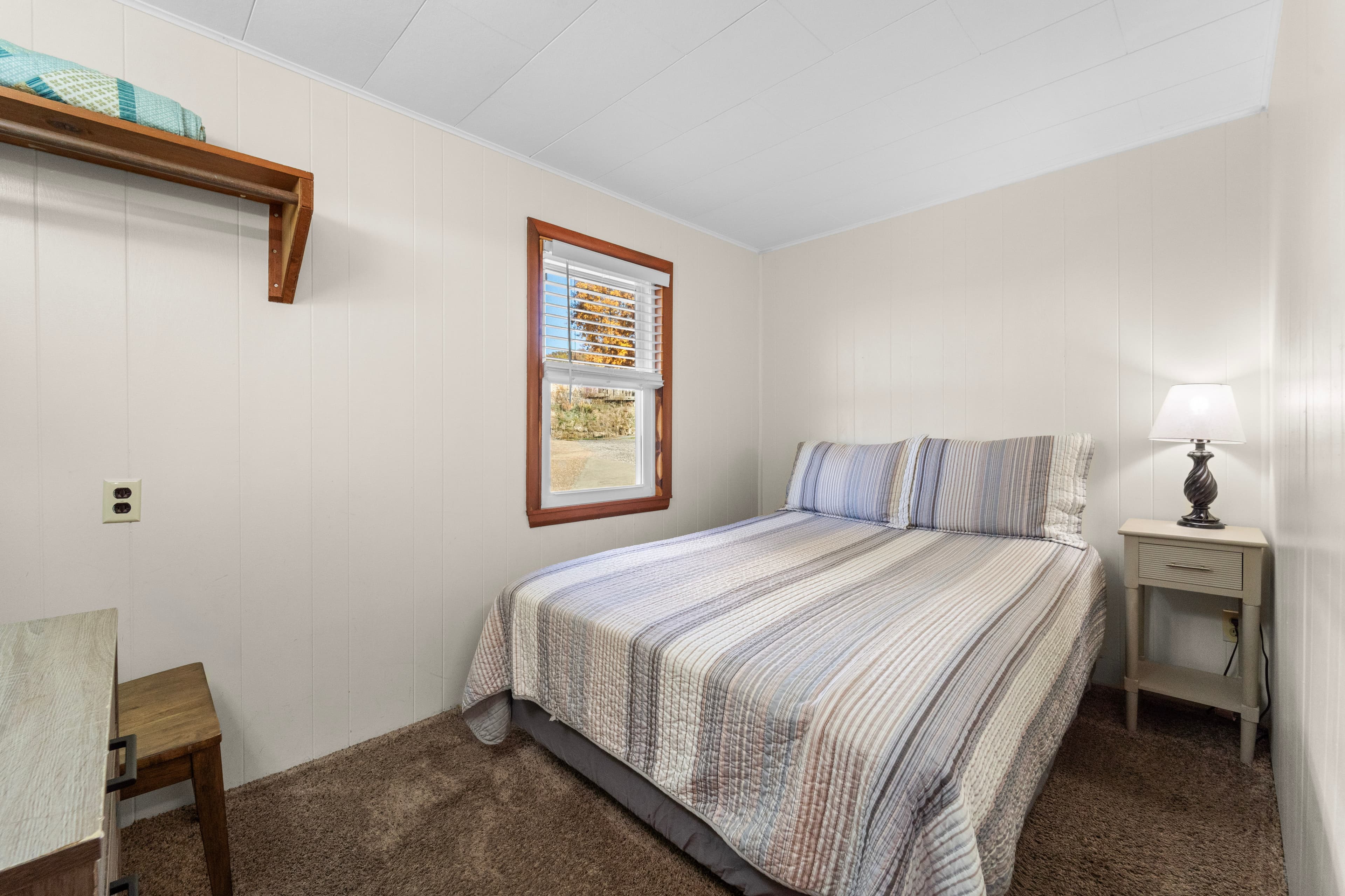 A cozy bedroom featuring a striped bedspread, a small nightstand with a lamp, and a window with natural light.