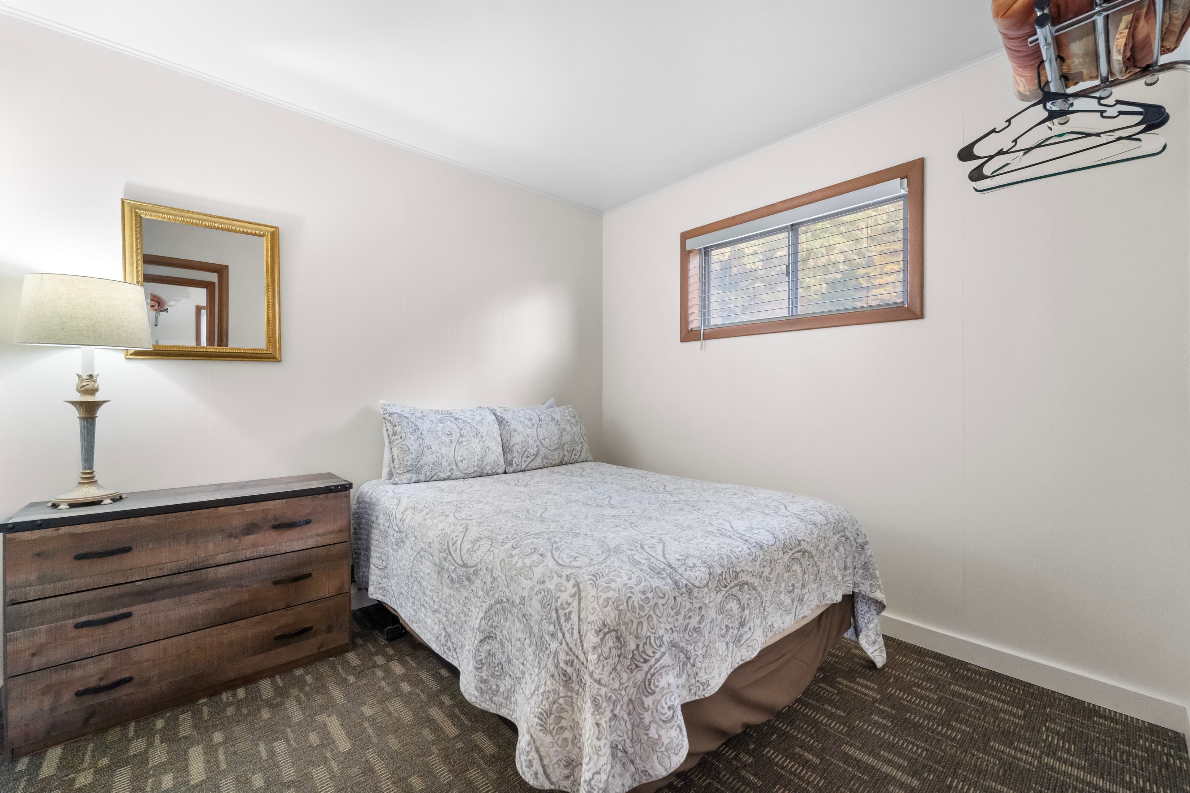 A cozy bedroom featuring a double bed, a wooden dresser, a lamp, and a window.