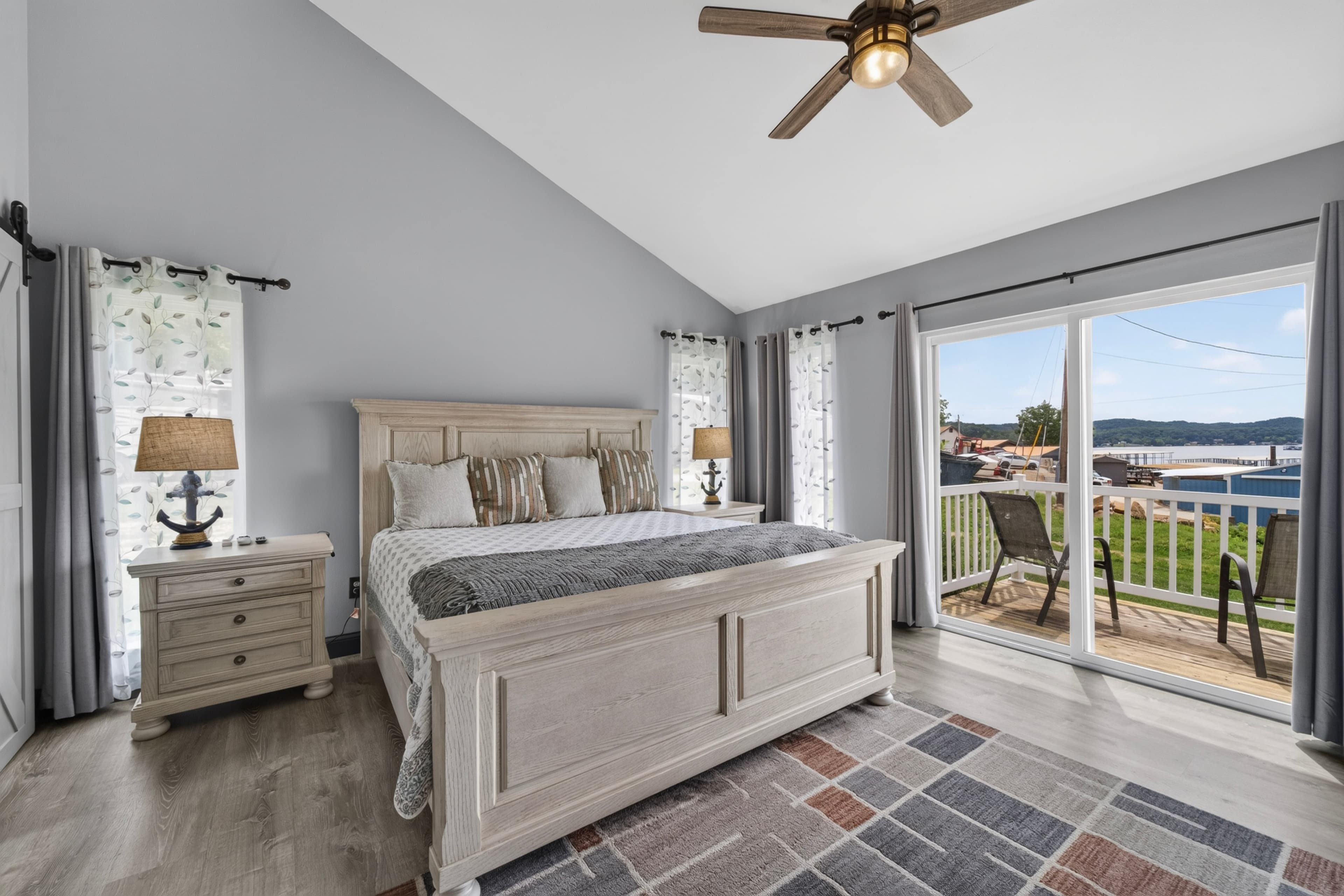 A cozy bedroom featuring a bed, nightstands, and large windows with a view of a deck and water outside.