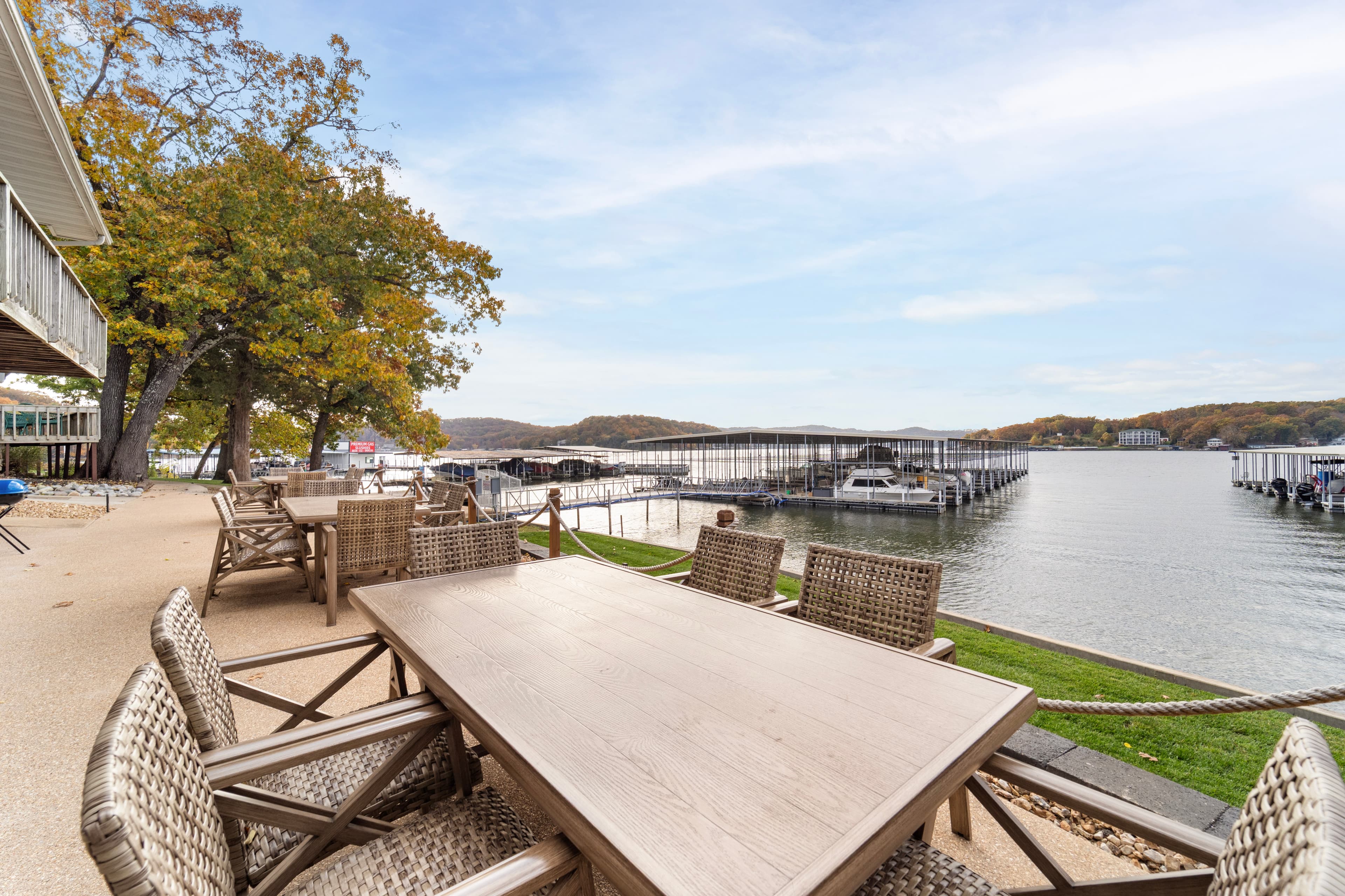A wooden dining table set on a patio overlooks a serene lake with boat docks and autumn trees in the background.