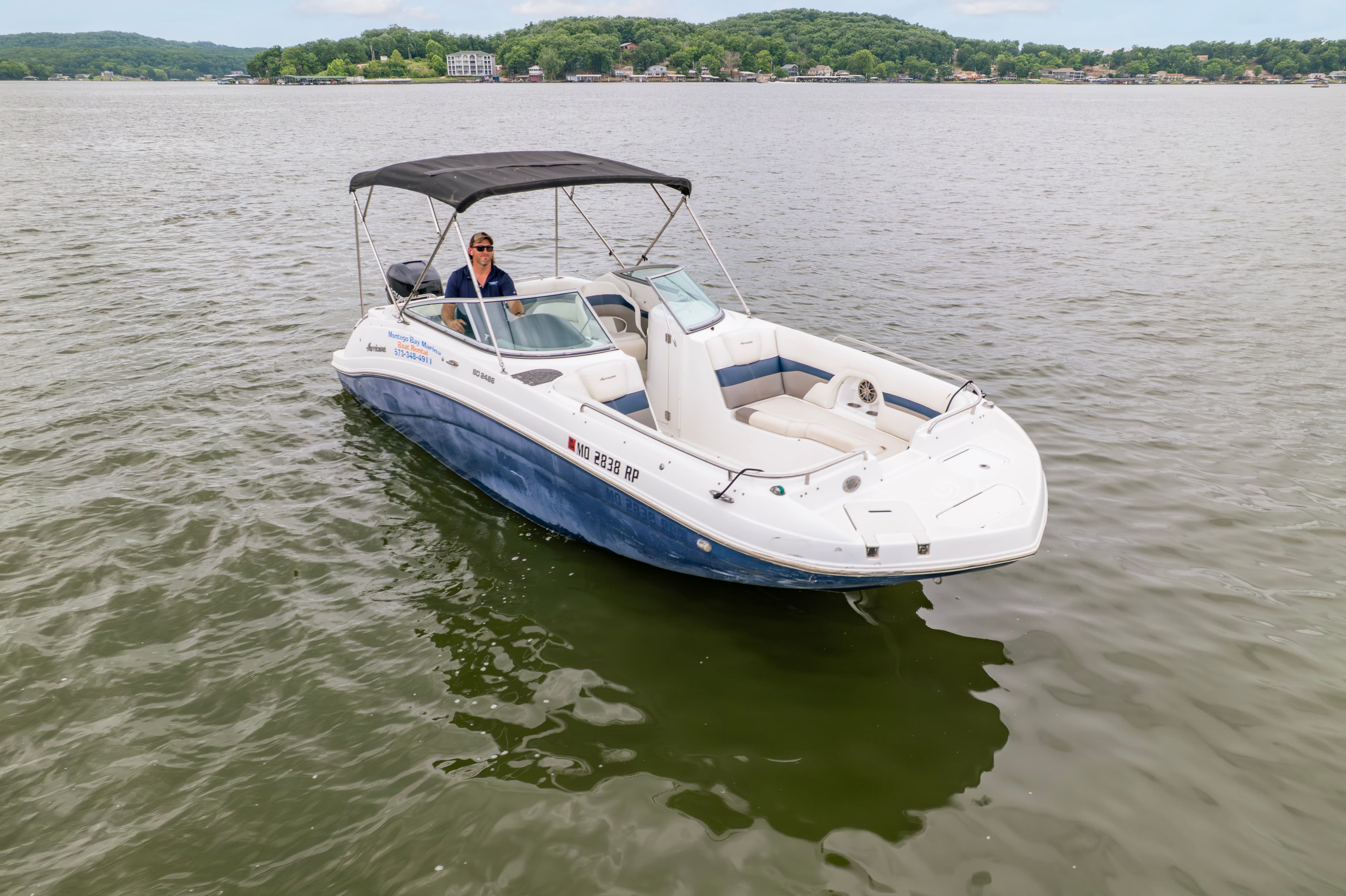 A person is seated at the helm of a white and blue boat on calm water, surrounded by green hills in the background.