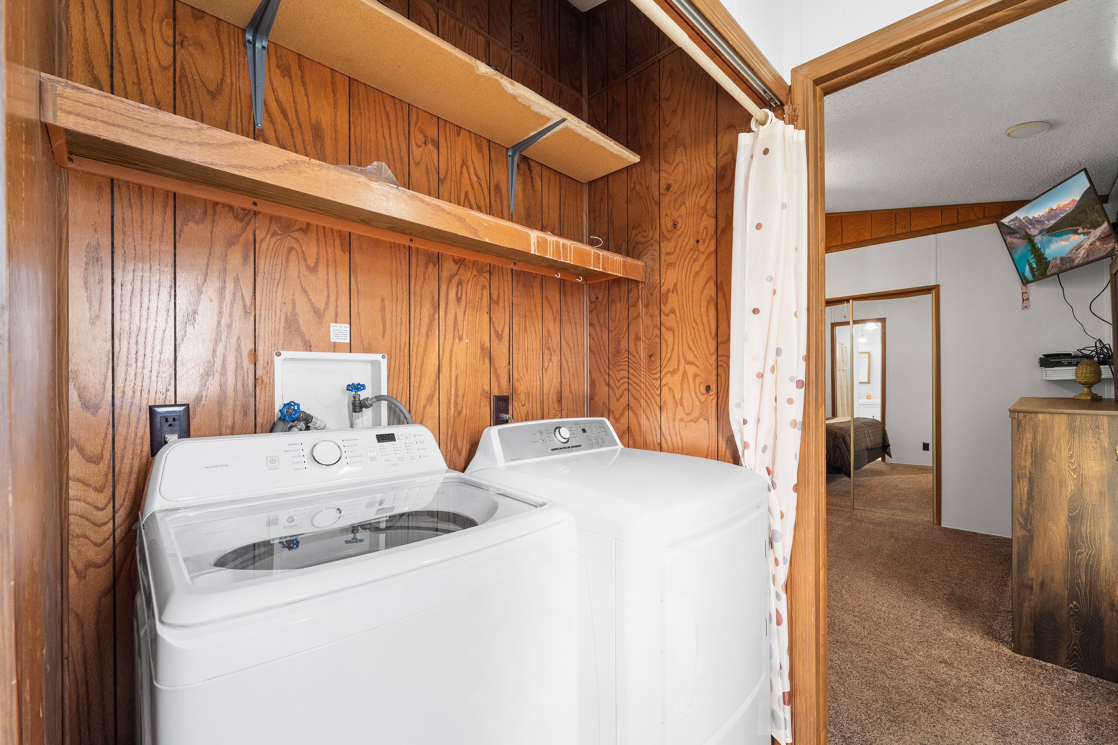 Laundry area with two machines and wooden paneling, leading to a bedroom.