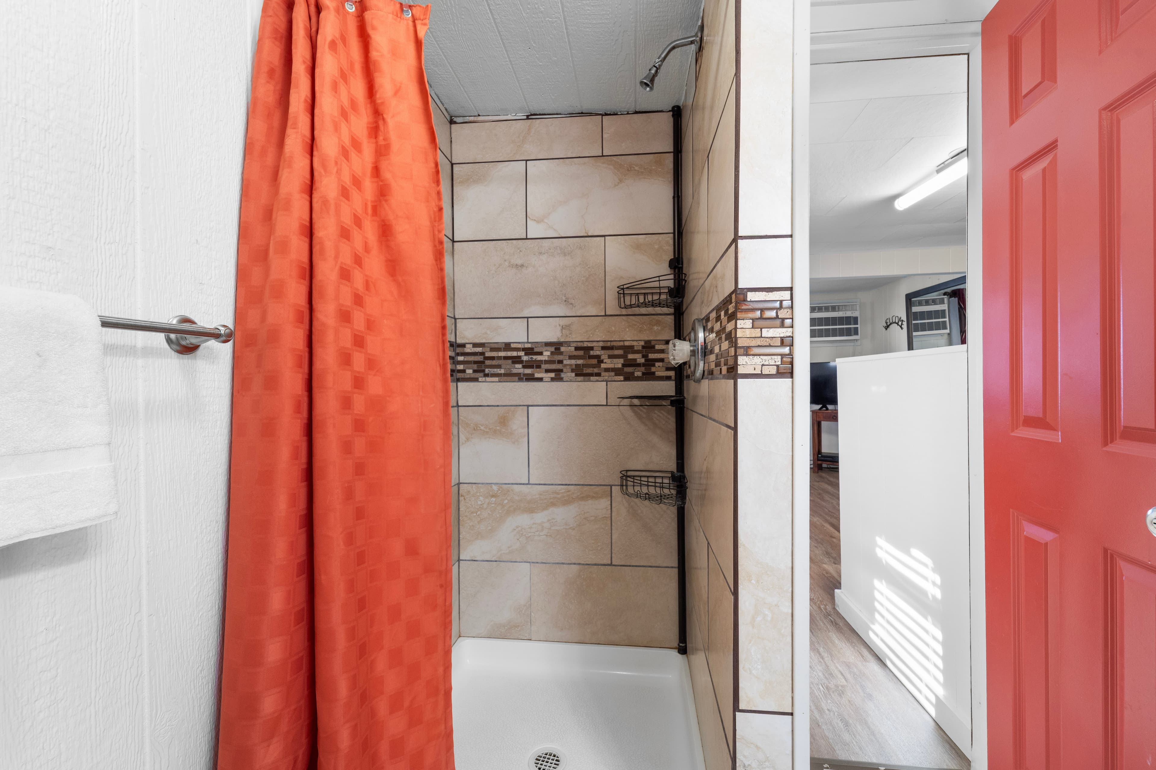 A shower with an orange curtain and tiled walls is shown, leading to a red door and a glimpse of an adjacent room.