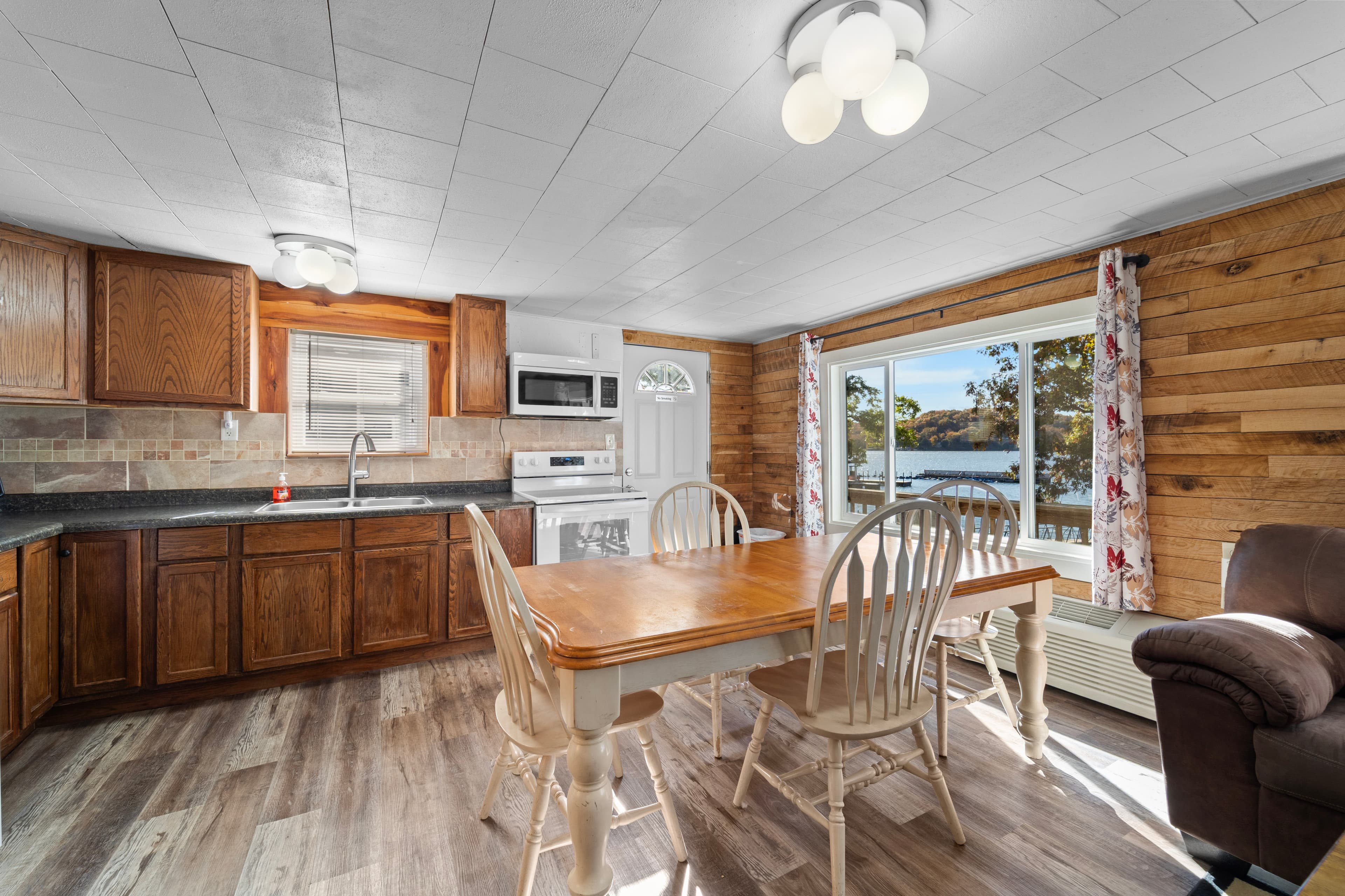 A bright, rustic kitchen featuring wooden cabinetry, a dining table, and a view of nature through a large window.