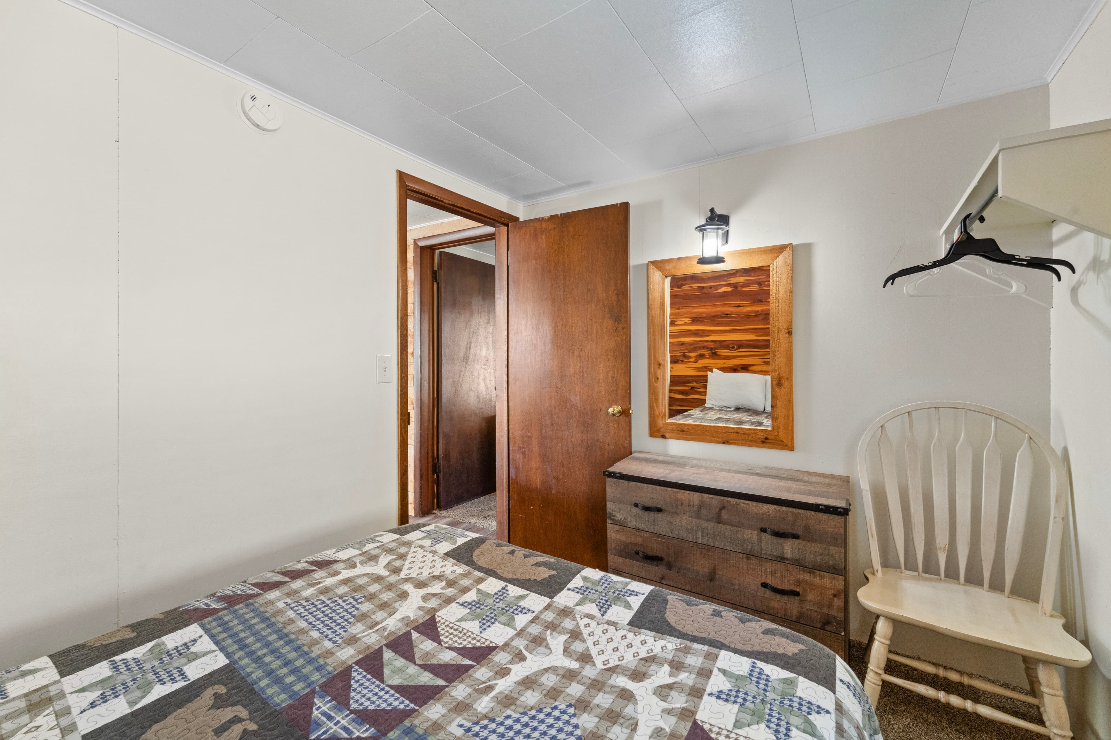 Cozy bedroom featuring a patterned quilt, wooden furniture, and a mirror with a light fixture.