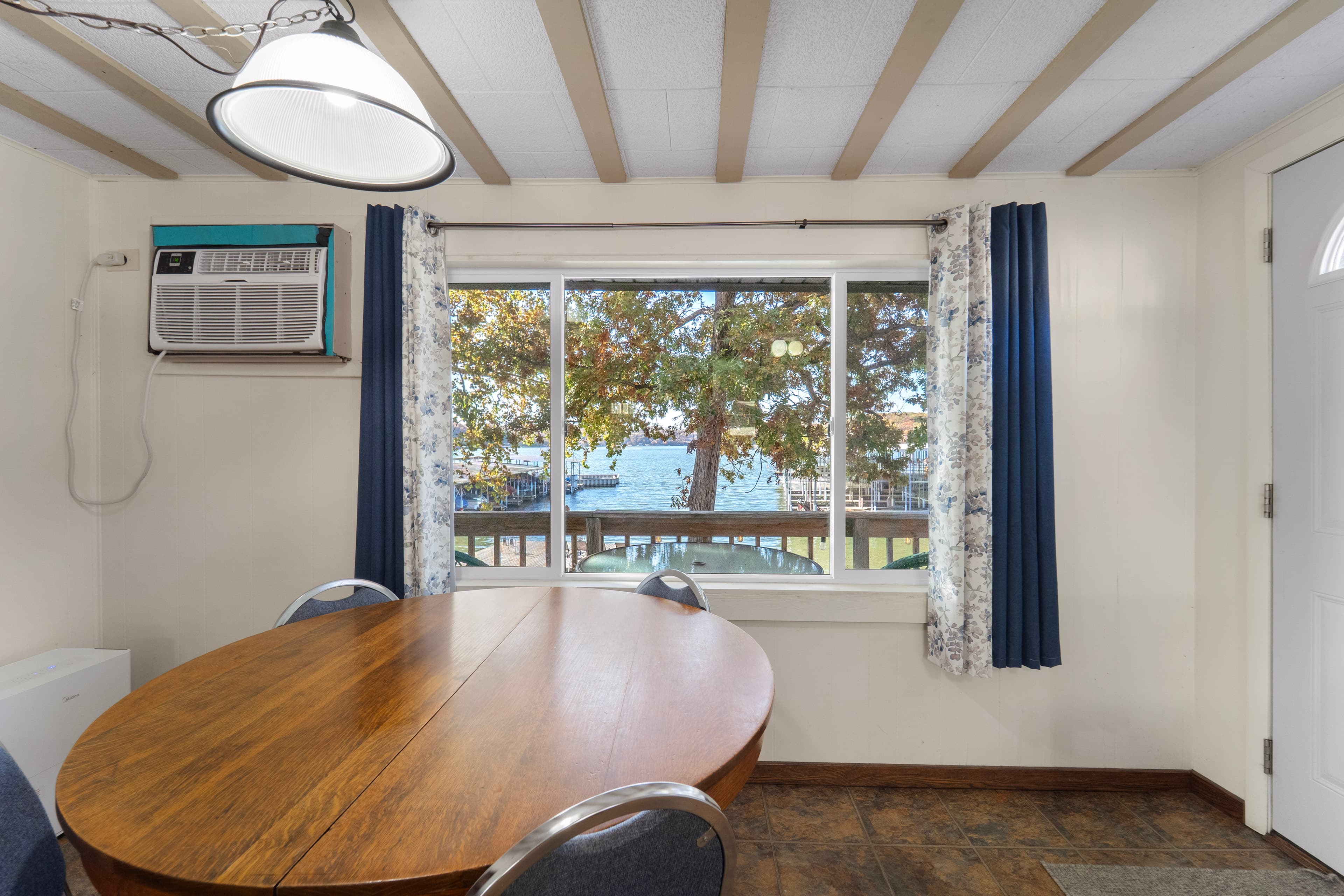 A cozy dining area features a wooden table, blue curtains, and a large window overlooking a lake.