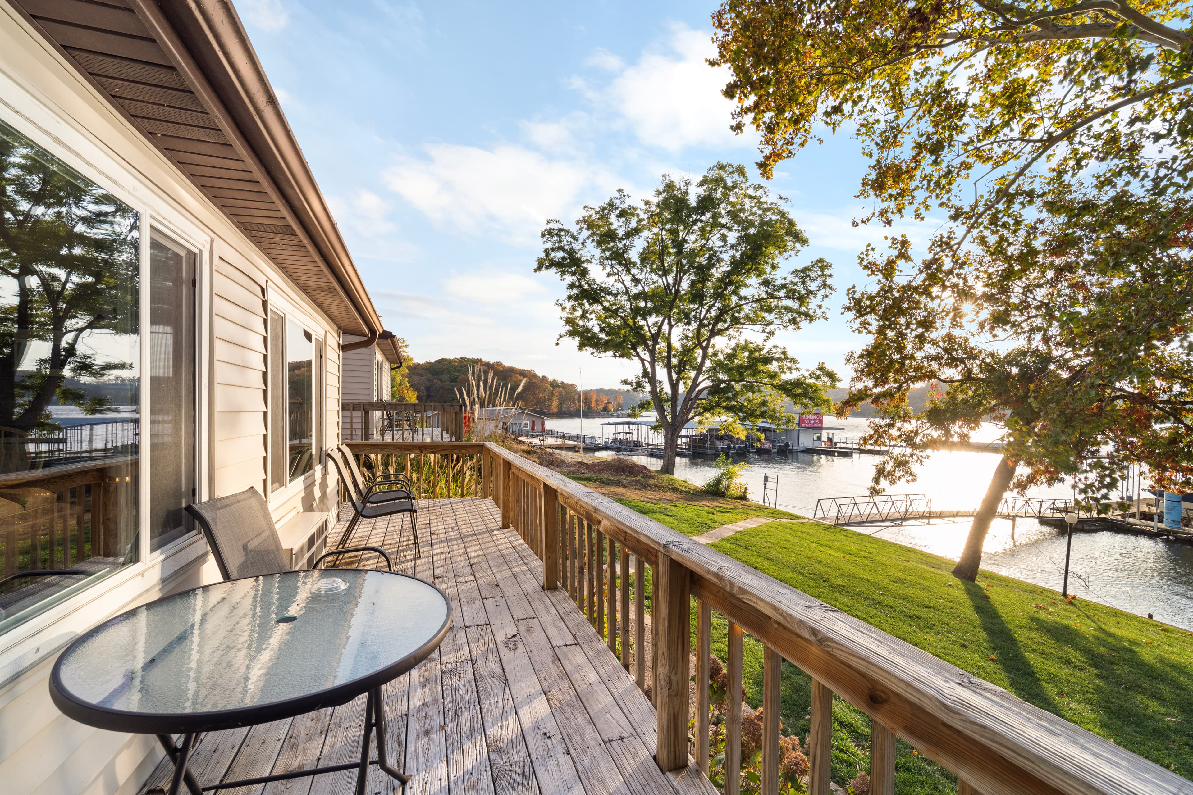 A wooden deck with a round table and chairs overlooks a serene waterway and trees under a blue sky.