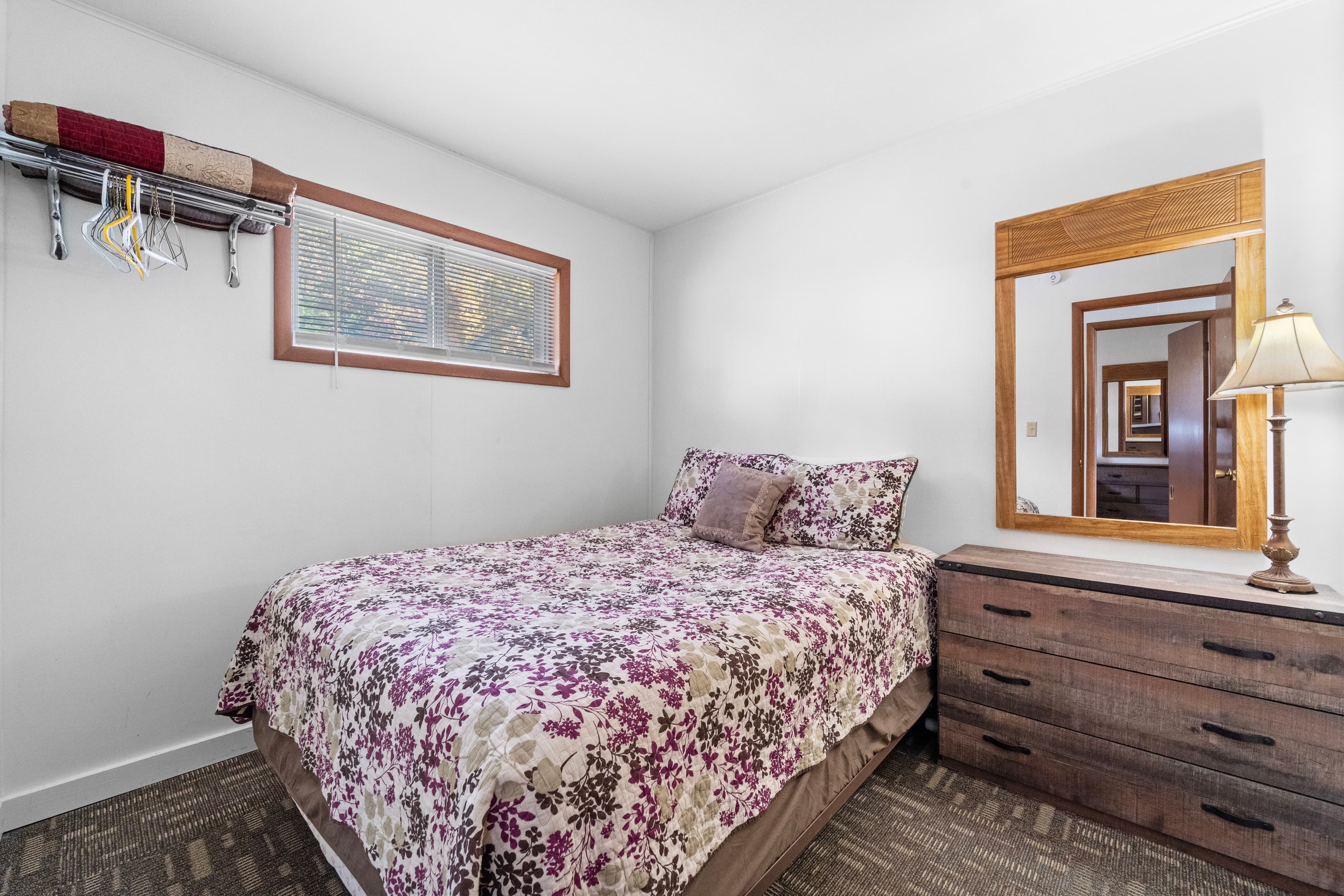 A cozy bedroom featuring a floral-patterned bedspread, wooden dresser, mirror, and natural light from a nearby window.