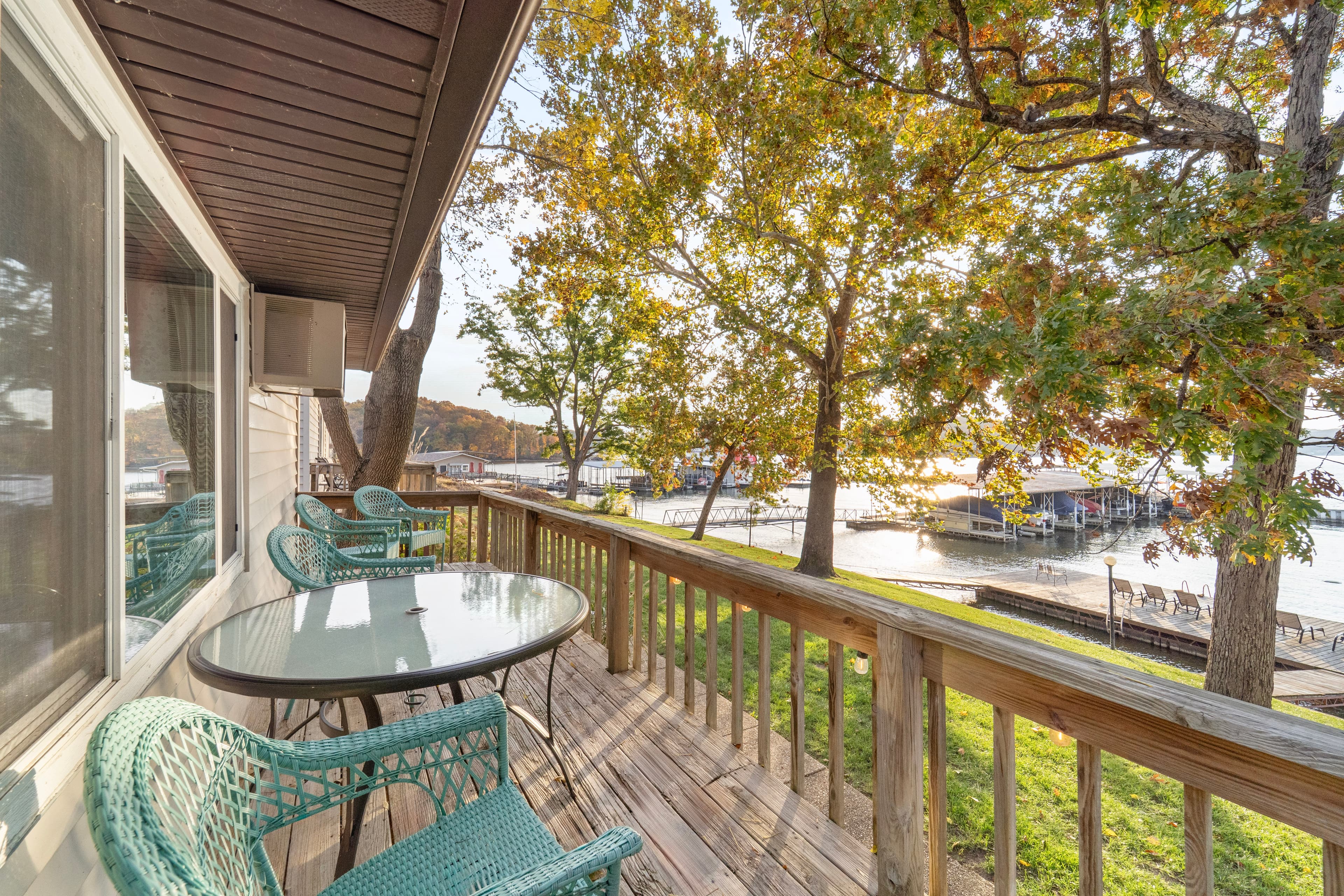 A scenic deck overlooking a tranquil lake, featuring a glass table and teal chairs surrounded by vibrant autumn foliage.