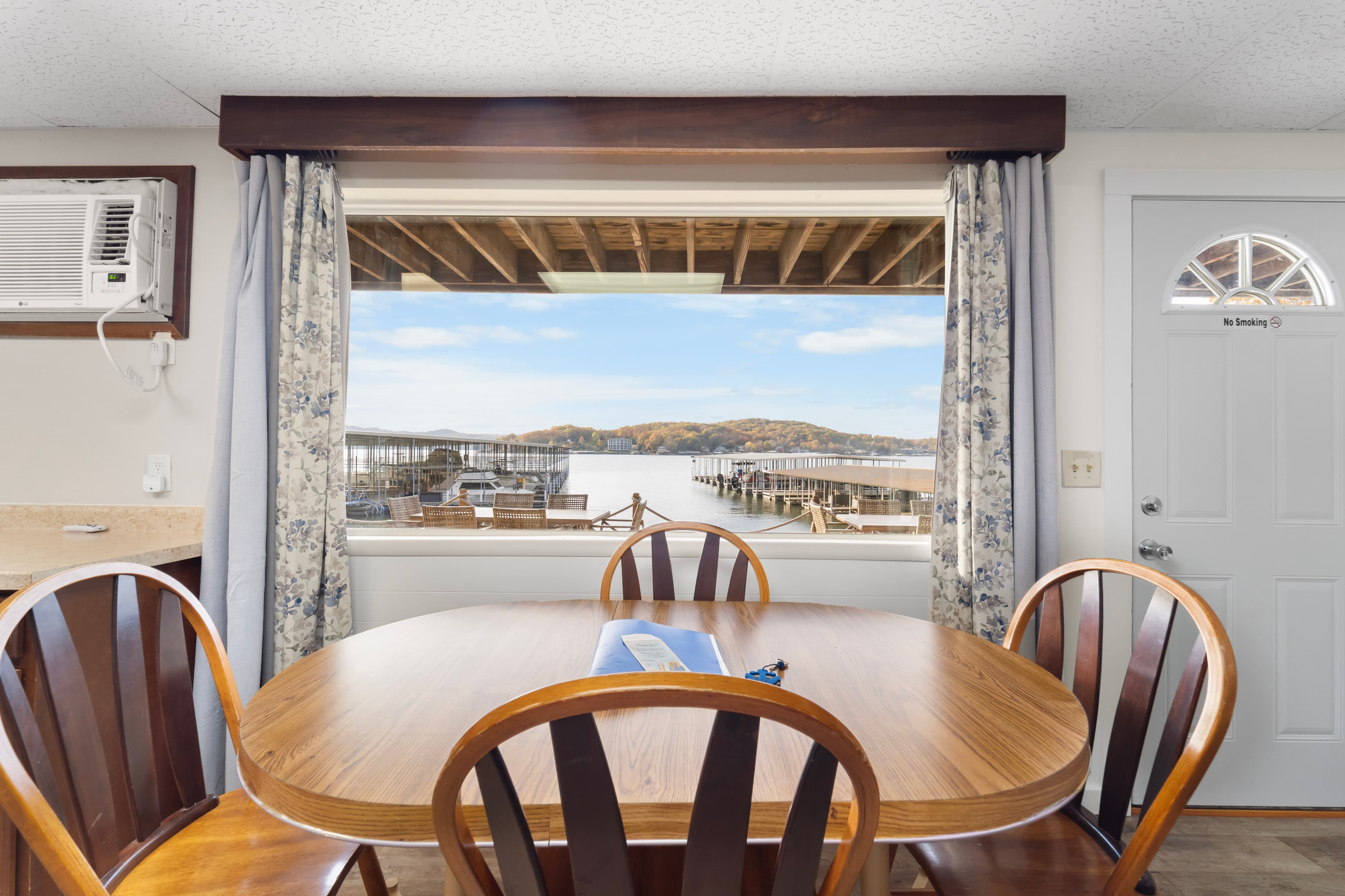 A dining table with wooden chairs sits in front of a large window showcasing a view of a lake and docks.