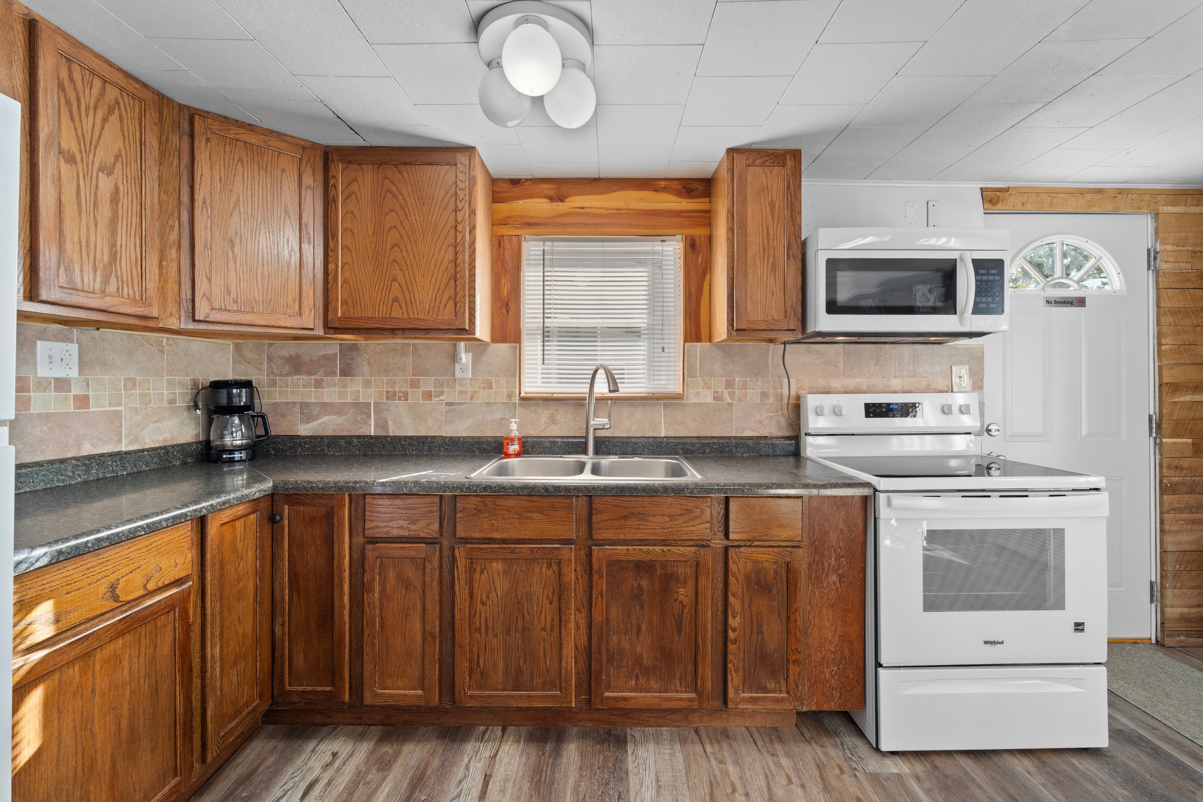 A modern kitchen featuring wooden cabinets, a double sink, a microwave, and a white stove with a door leading outside.