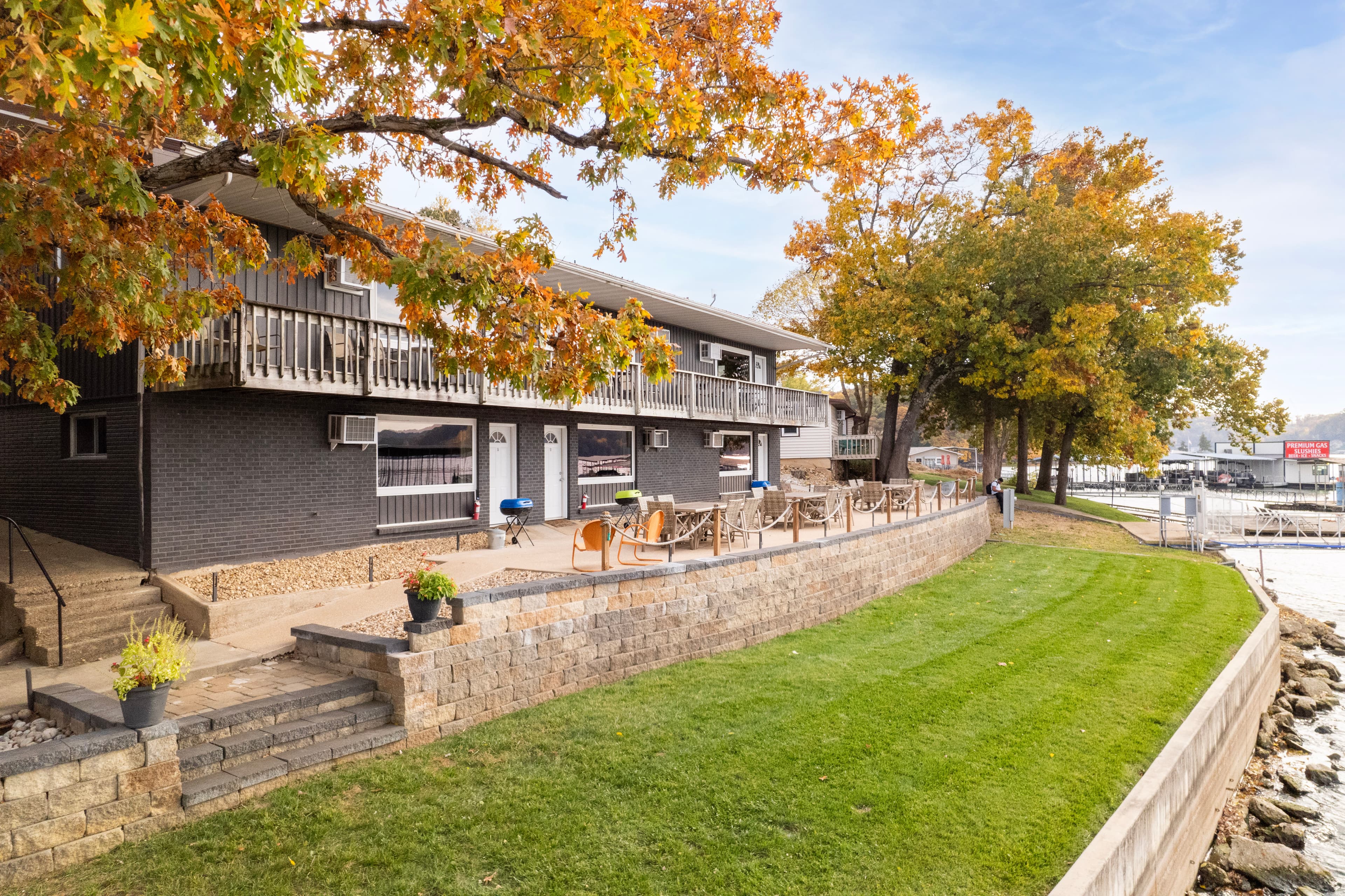 A lakeside property with autumn foliage and a grassy area, featuring a patio and seating near the water.