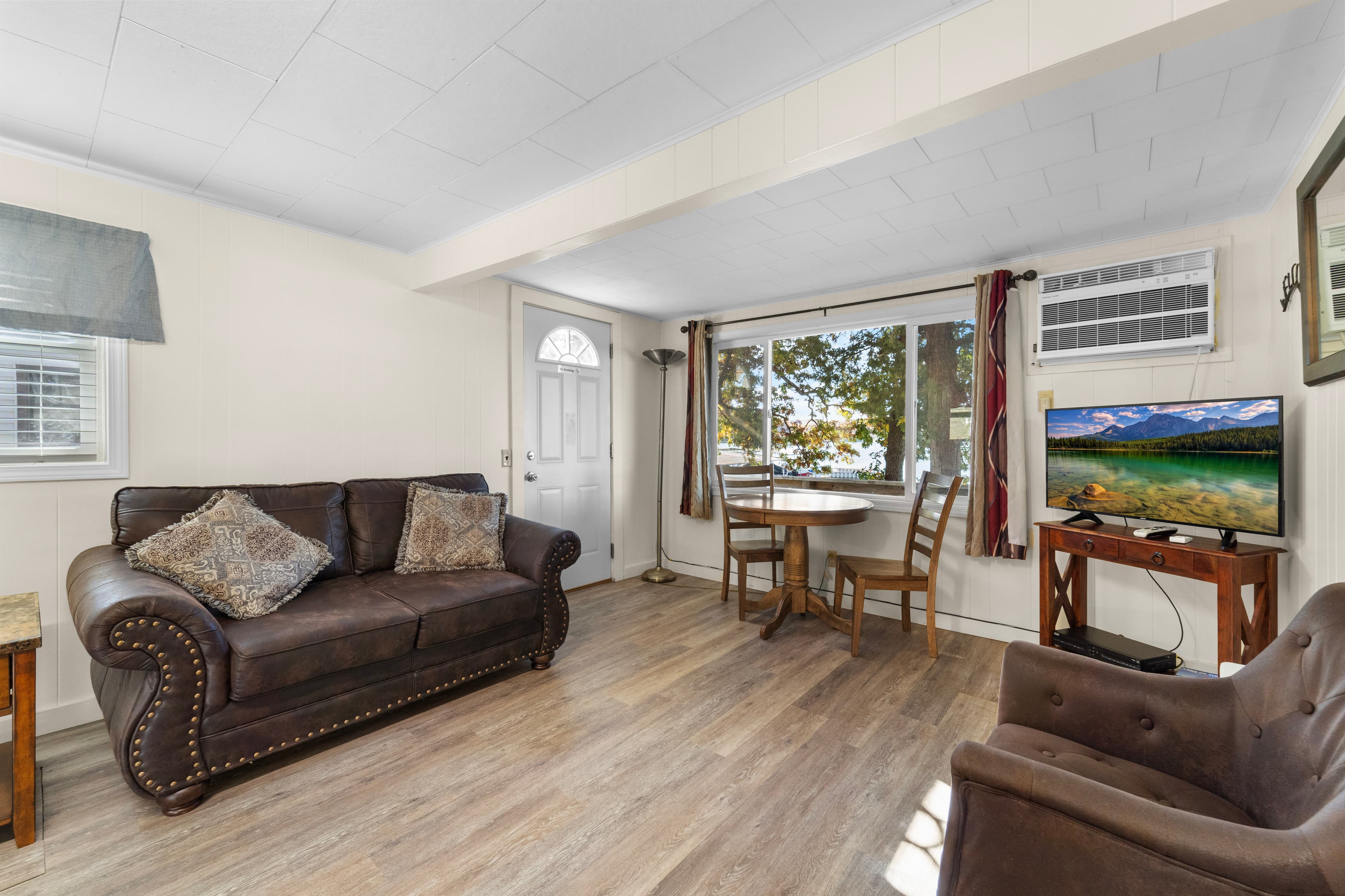 A cozy living room featuring a dark leather sofa, a dining table by a large window, and a TV.