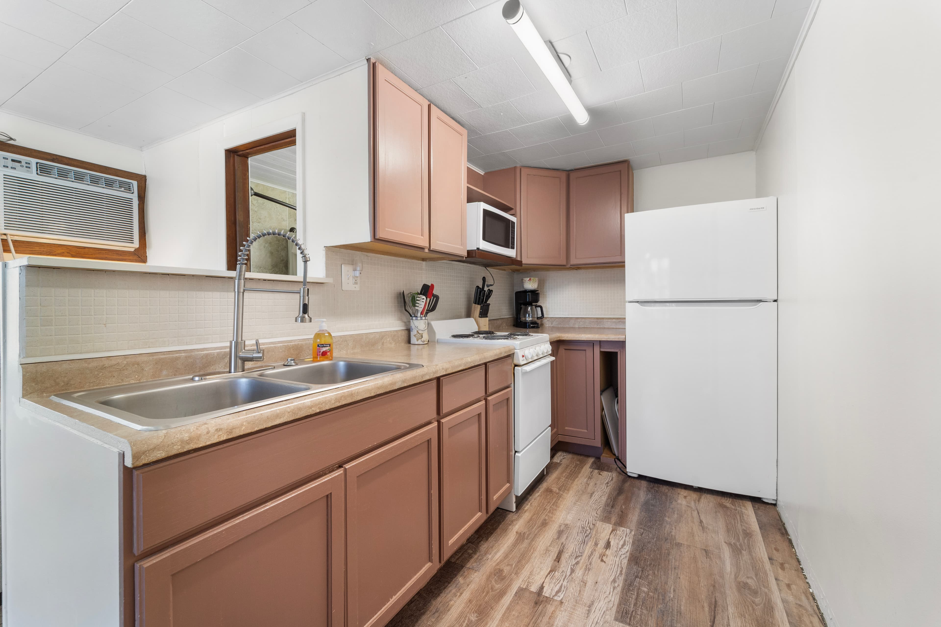A compact kitchen featuring brown cabinetry, a double sink, white appliances, and wood flooring.