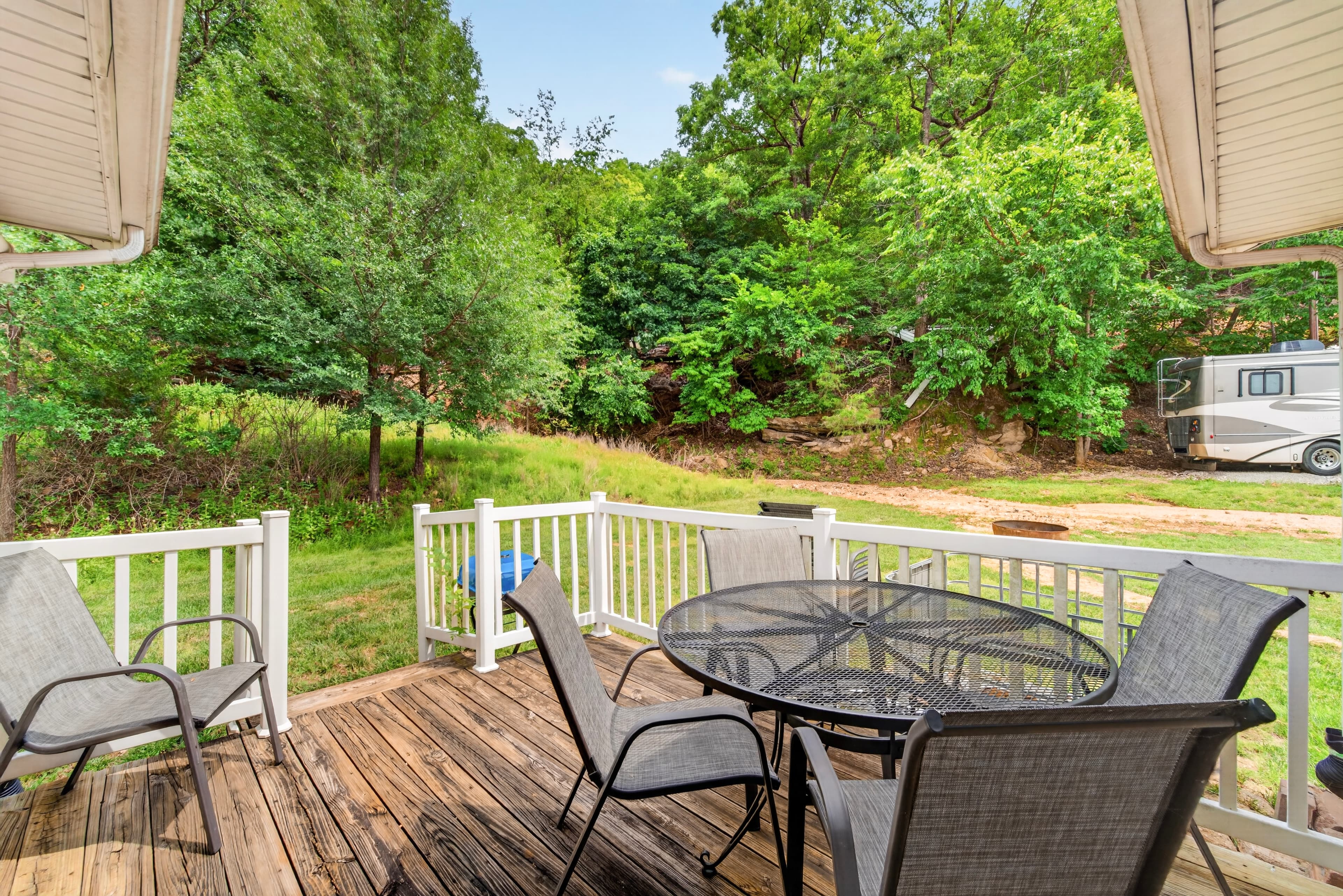 A wooden deck with a round table and chairs overlooks a green, wooded area.