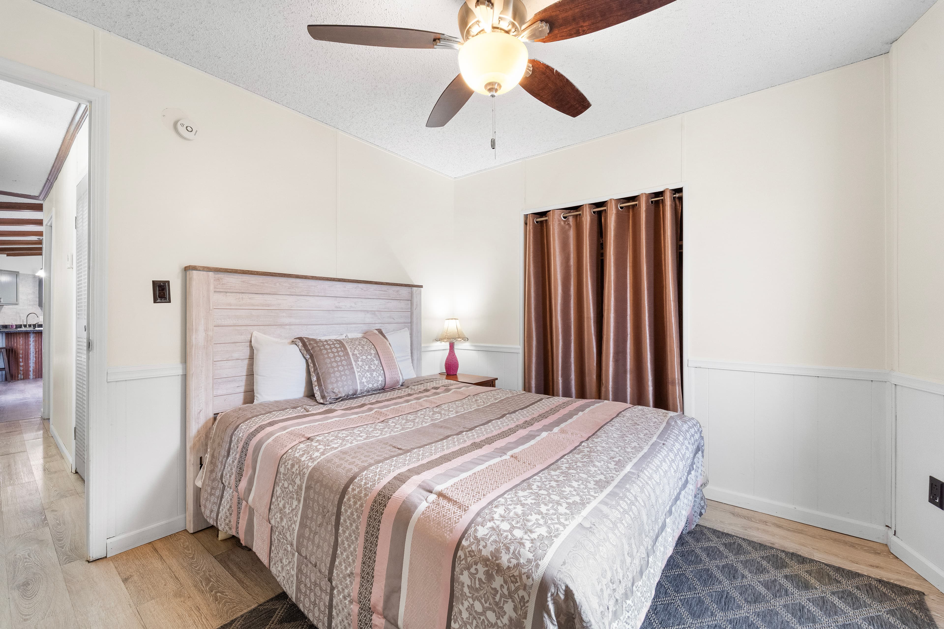 A cozy bedroom featuring a double bed with patterned bedding, a ceiling fan, and a window with brown curtains.