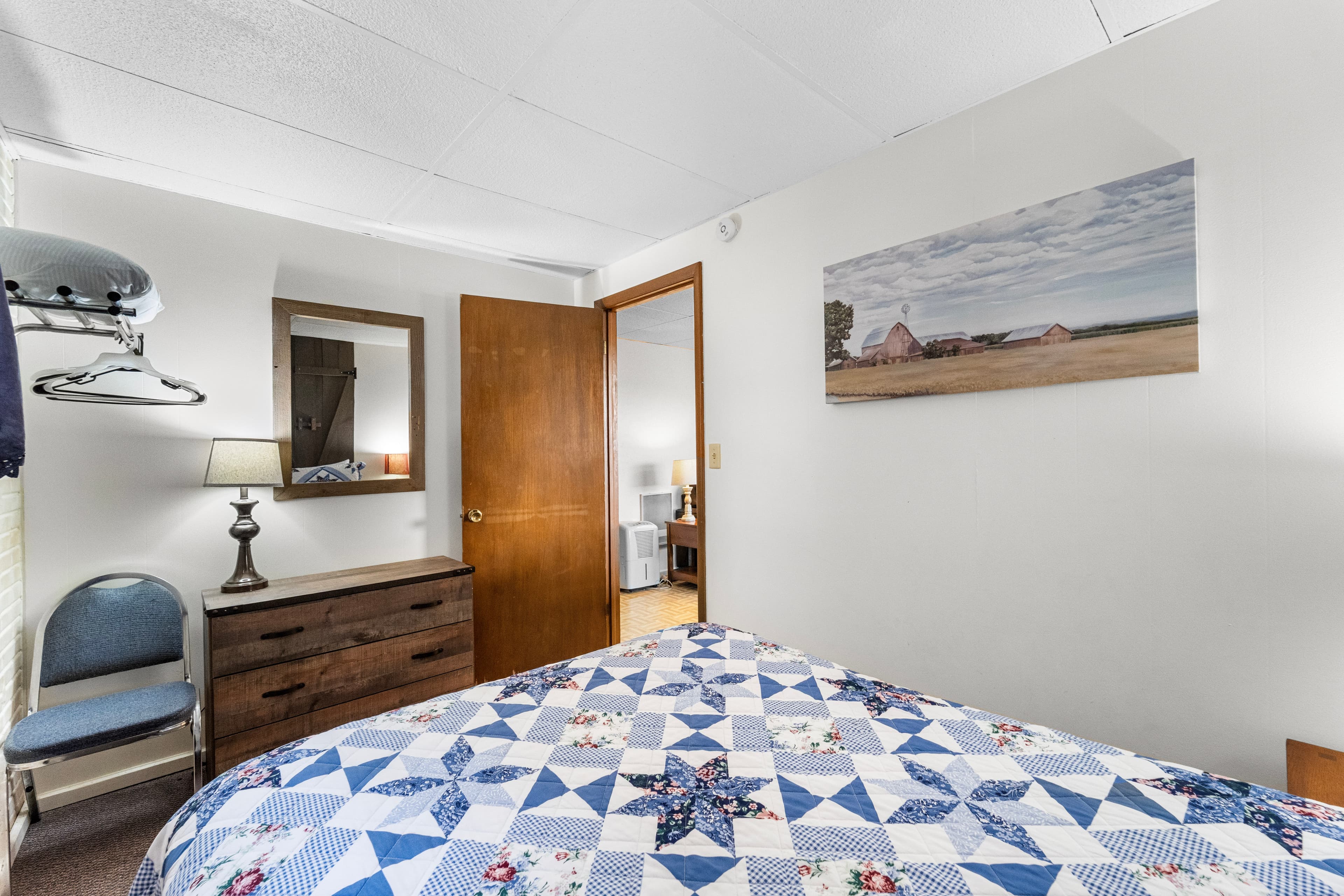 A cozy bedroom featuring a patchwork quilt, a dresser with a mirror, and a view of the door and a landscape painting on the wall.