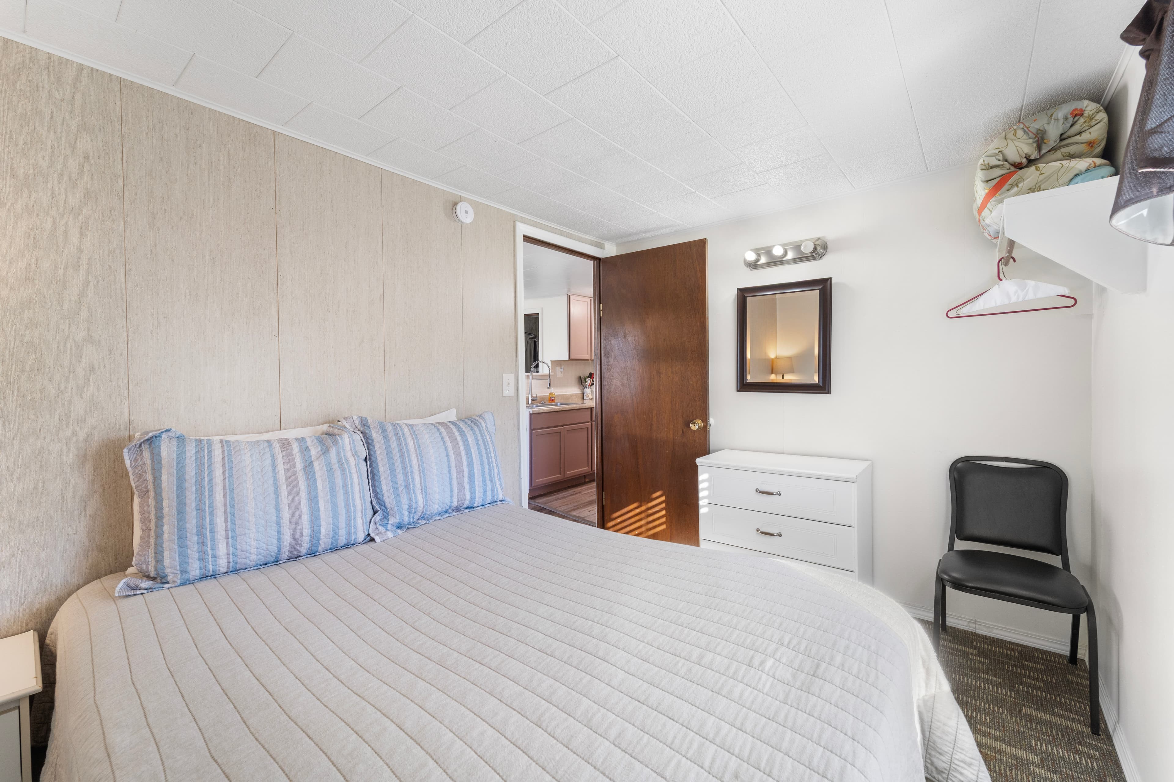 A bright, minimalistic bedroom featuring a double bed, dresser, and a door leading to a kitchen area.