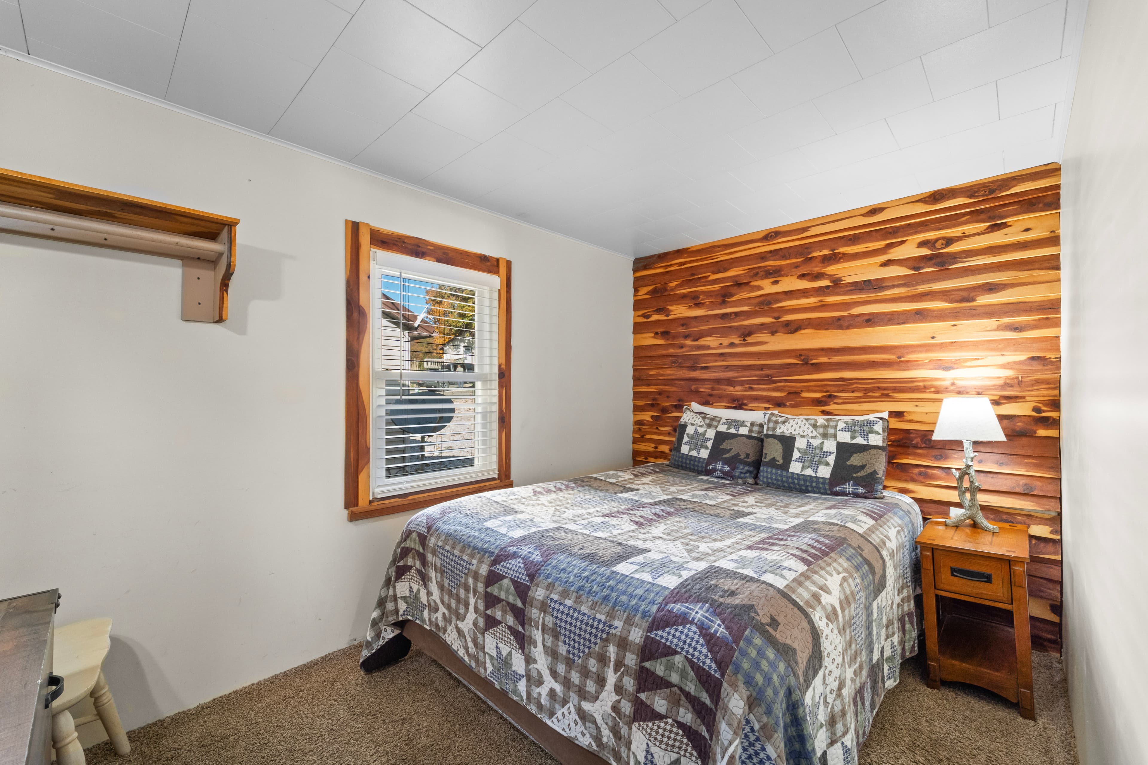 Cozy bedroom featuring a bed with a patterned quilt, wooden accent wall, and a bedside lamp.