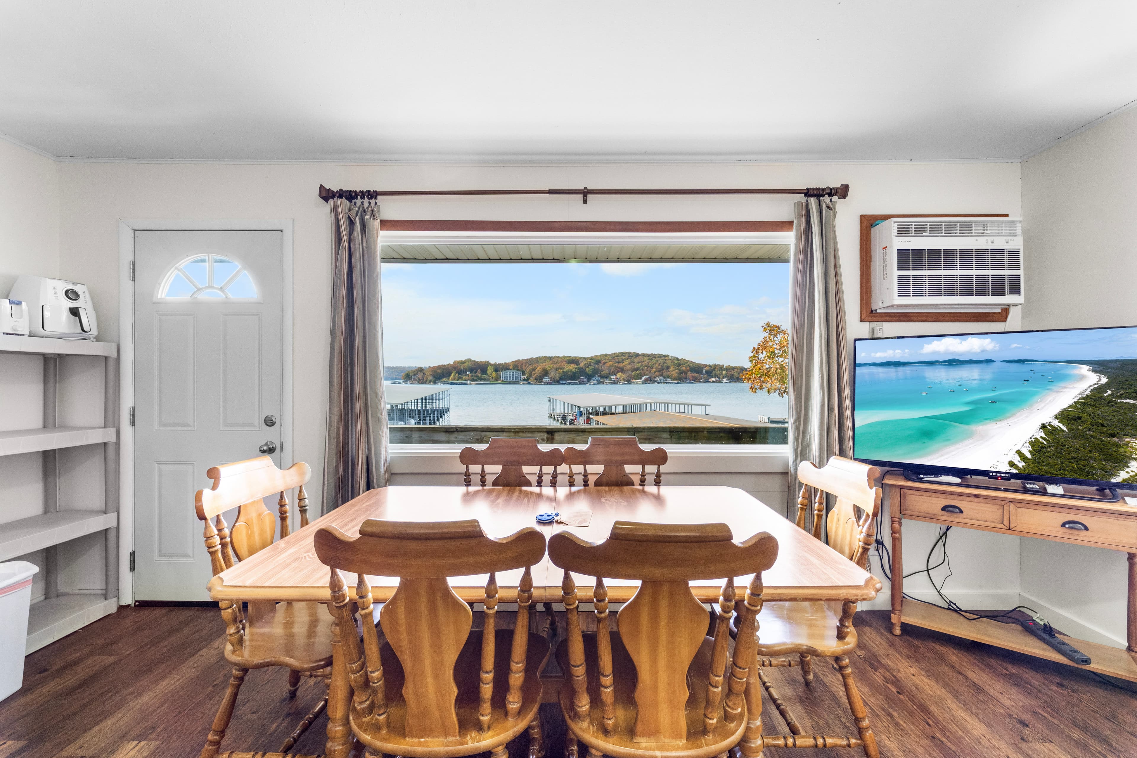 A dining area with wooden chairs and a table overlooks a scenic waterfront through a large window.