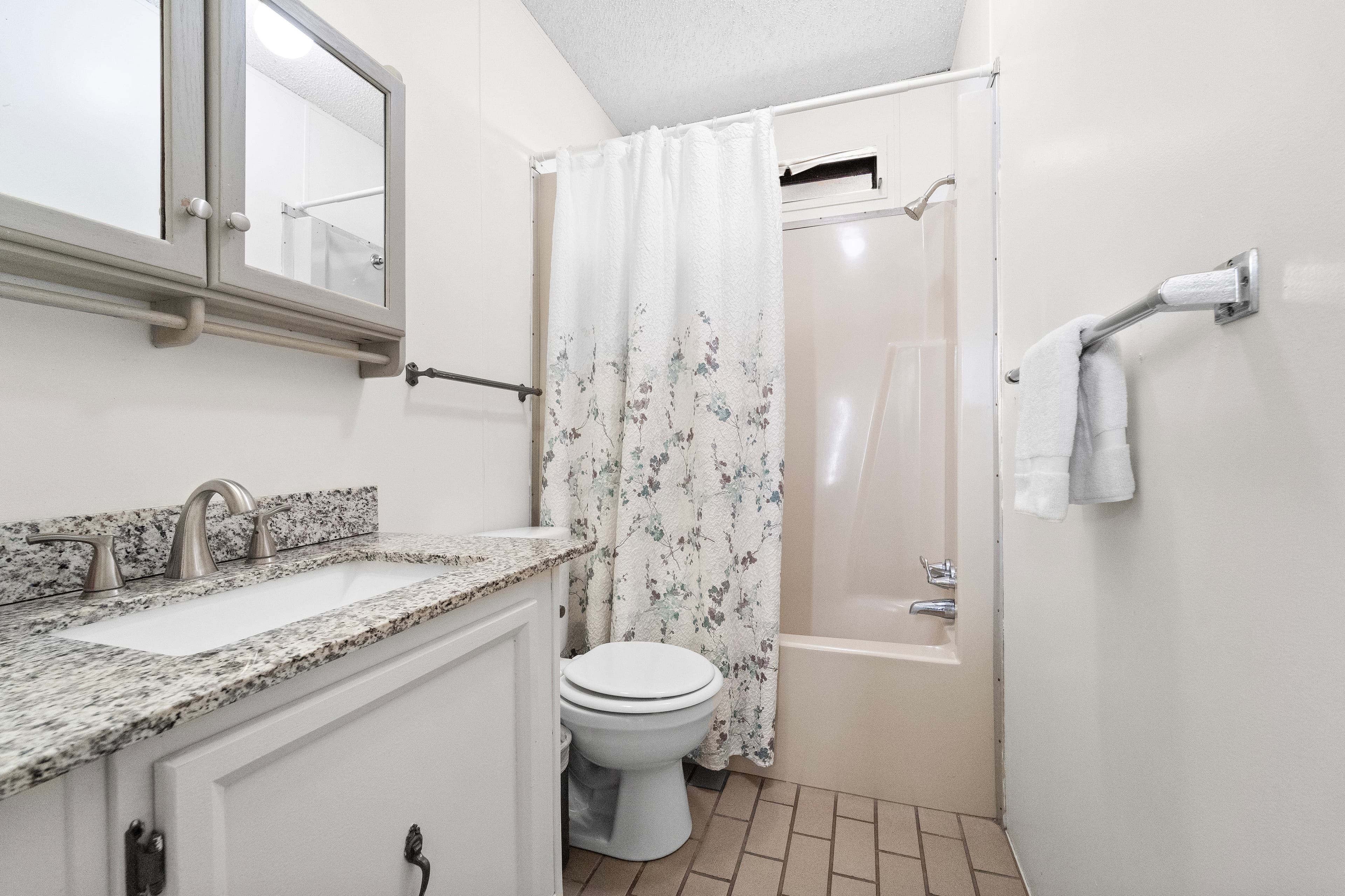 A compact bathroom featuring a granite countertop sink, a toilet, and a shower with a floral curtain.