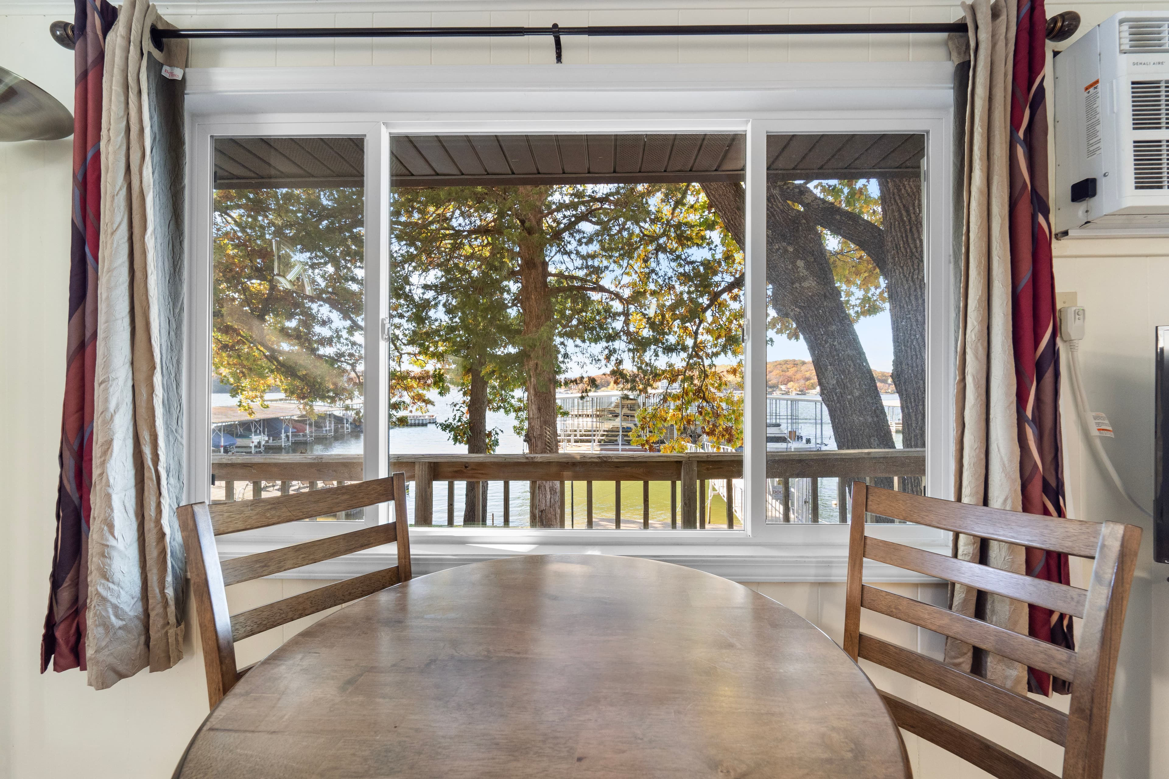 A wooden table and two chairs sit by a window overlooking trees and a lakefront.