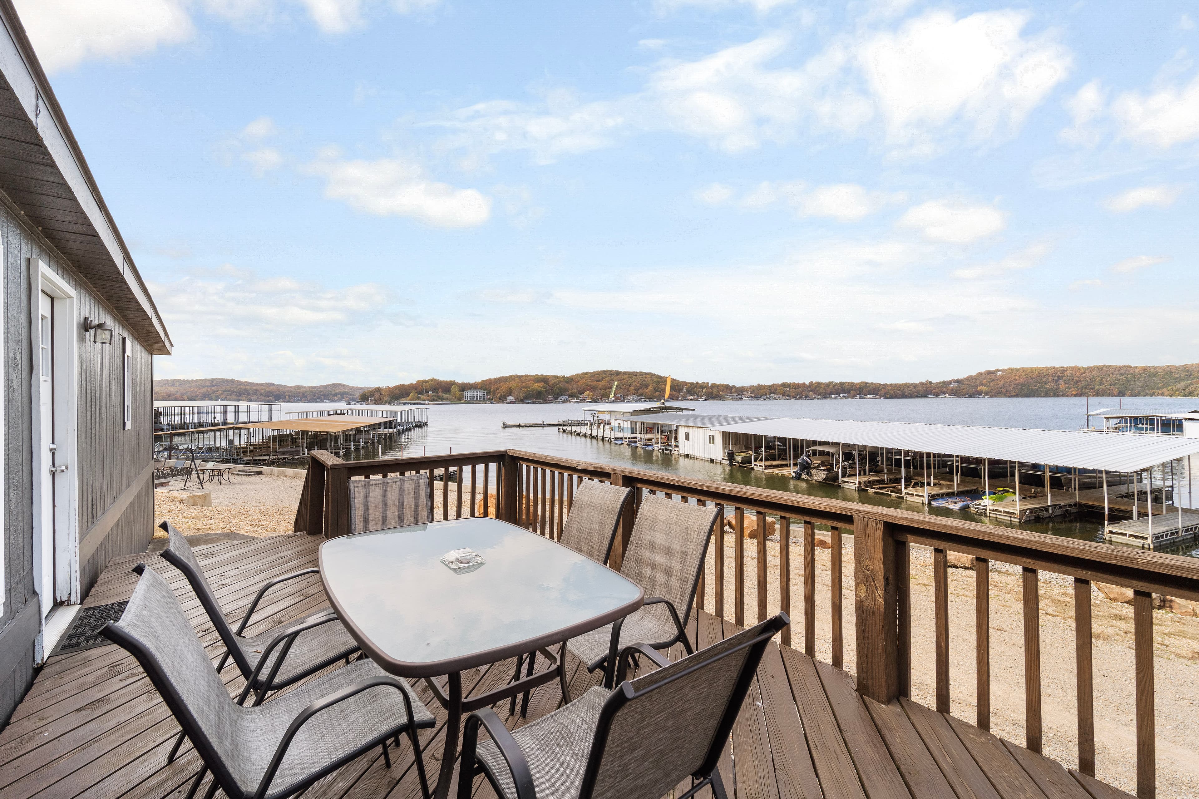 A wooden deck with table and chairs overlooks a tranquil lake with marina docks in the background.