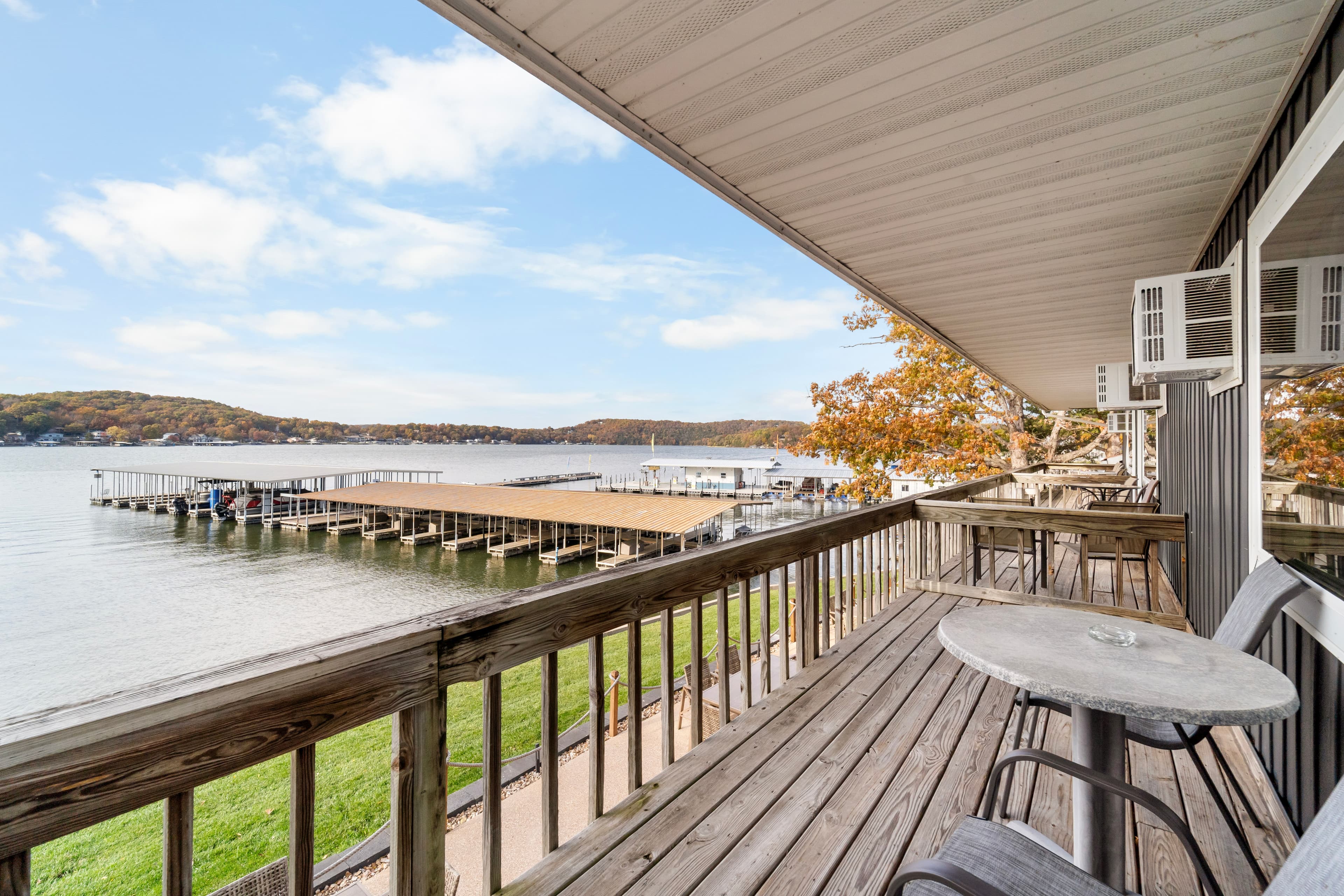 A wooden balcony overlooking a serene lake with docks, surrounded by autumn foliage and a clear blue sky.