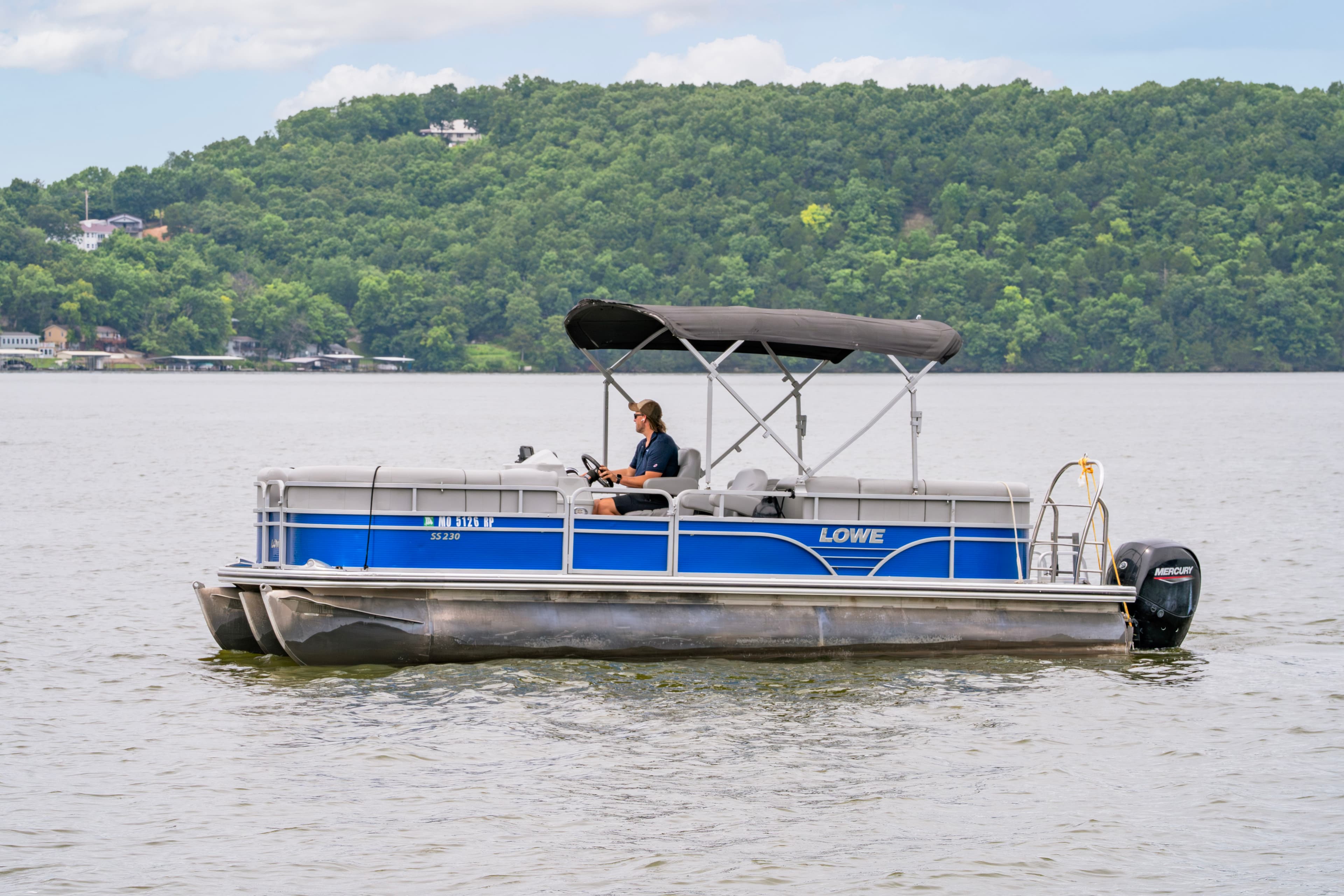 A person is steering a blue pontoon boat on a lake with lush green hills in the background.