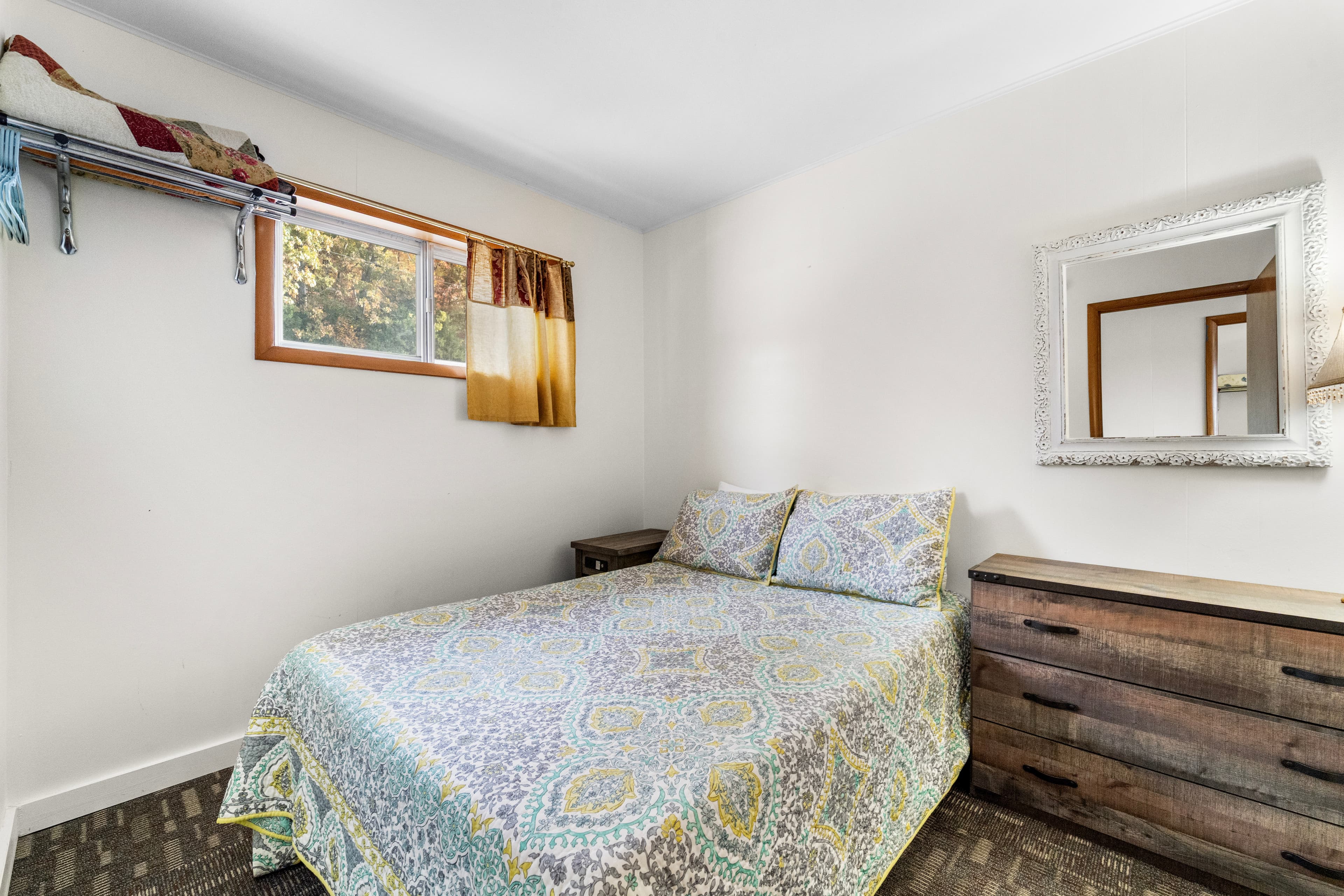 A cozy bedroom featuring a double bed with a colorful patterned quilt, a wooden dresser, and a window with light drapes.