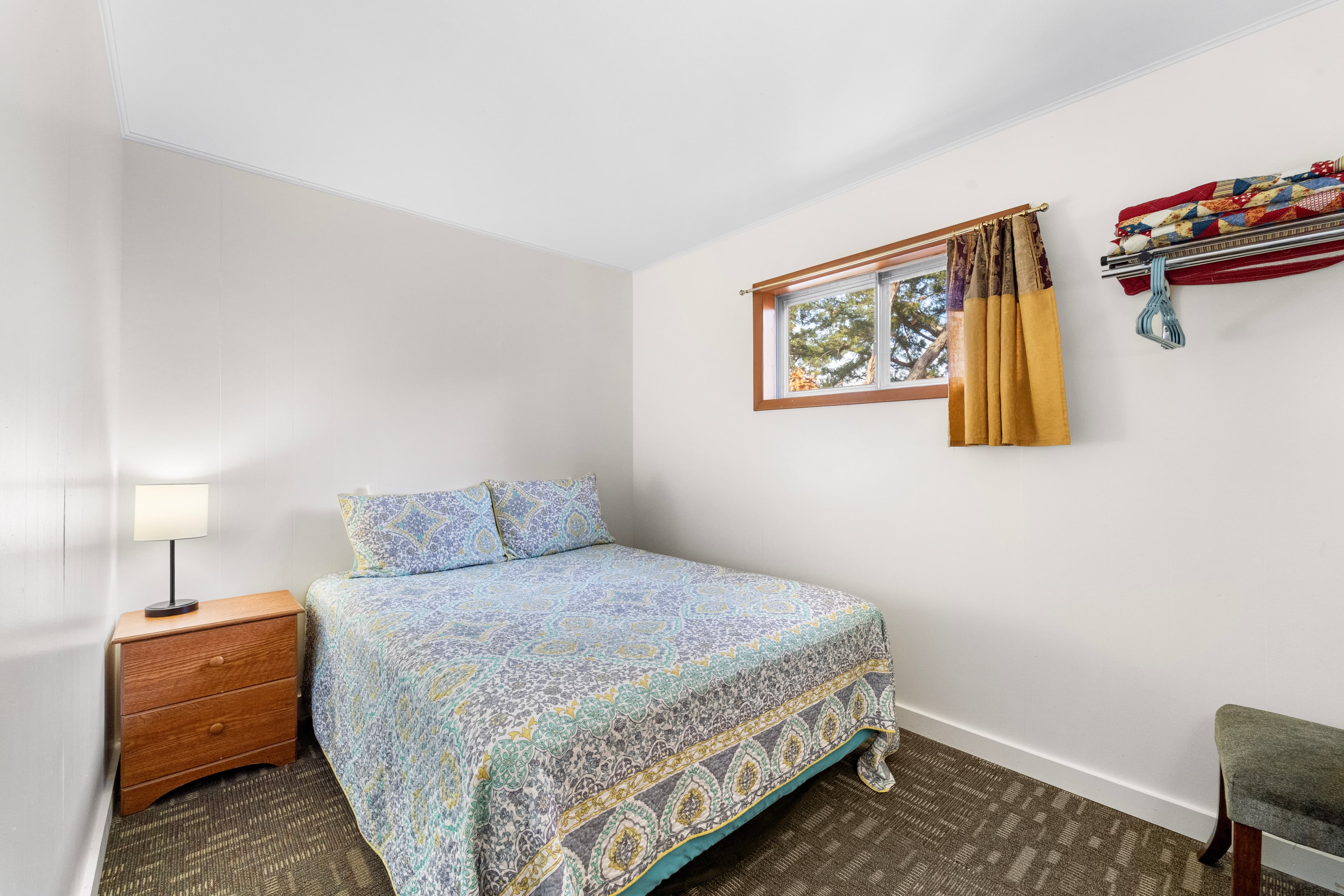 Cozy bedroom featuring a bed with a patterned quilt, a nightstand with a lamp, and a window with colorful curtains.
