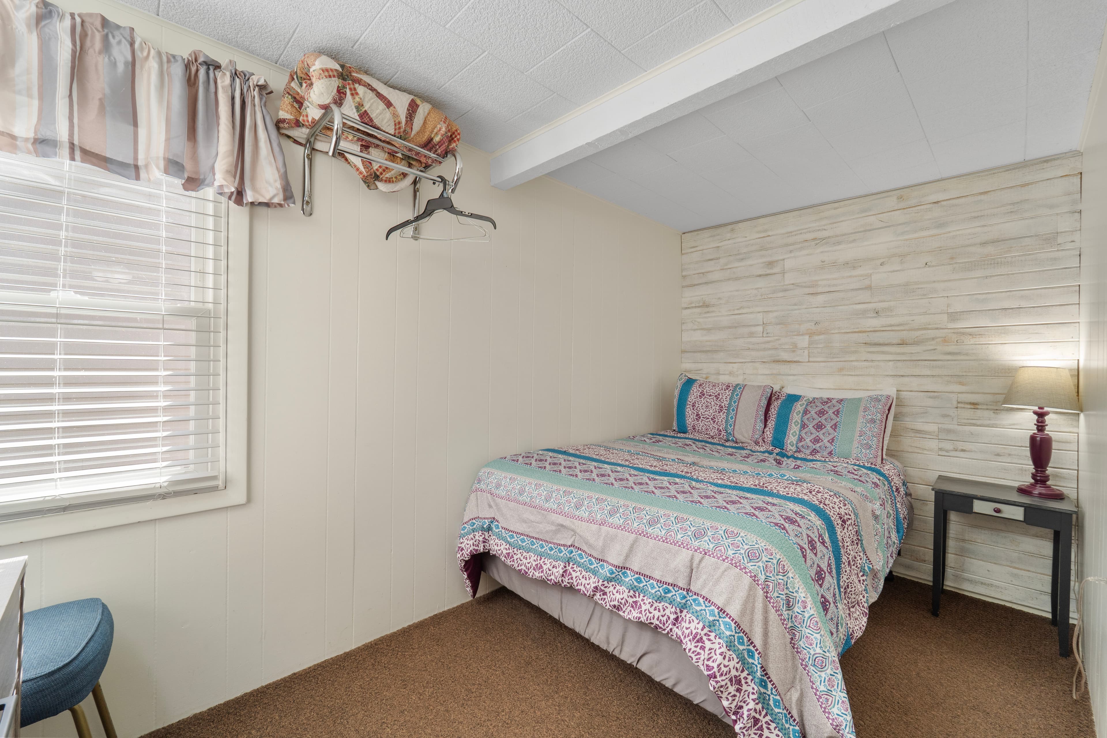 Cozy bedroom featuring a patterned bedspread, a bedside lamp, and a small chair next to a window.
